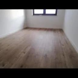 Jasne panele podłogowe z widoczną strukturą drewna w pustym pokoju z białymi ścianami i oknem w tle.