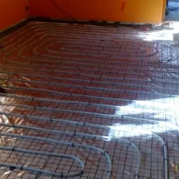 Instalacja ogrzewania podłogowego z rur rozprowadzonych na siatce w pomieszczeniu w trakcie remontu, pomarańczowe ściany w tle.