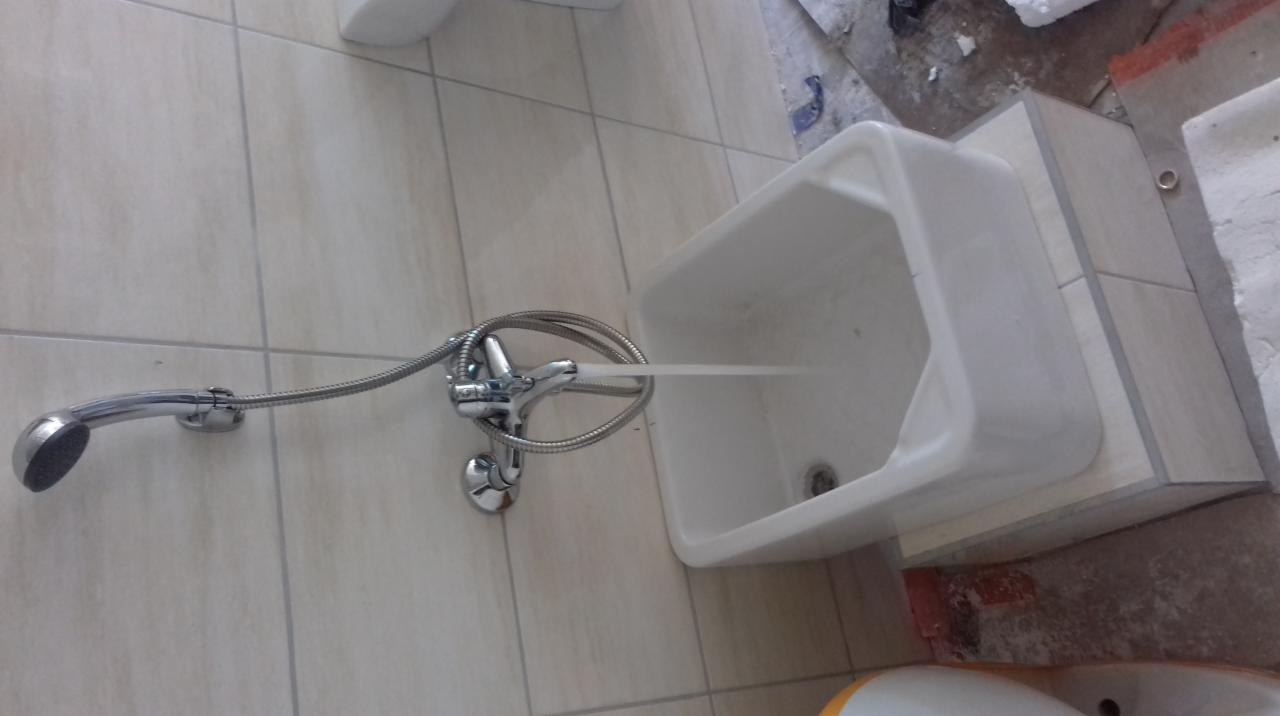 Biała umywalka z baterią i wężem prysznicowym zamontowana na ścianie z jasnych płytek, woda leje się z kranu. Widoczne ślady prac wykończeniowych.