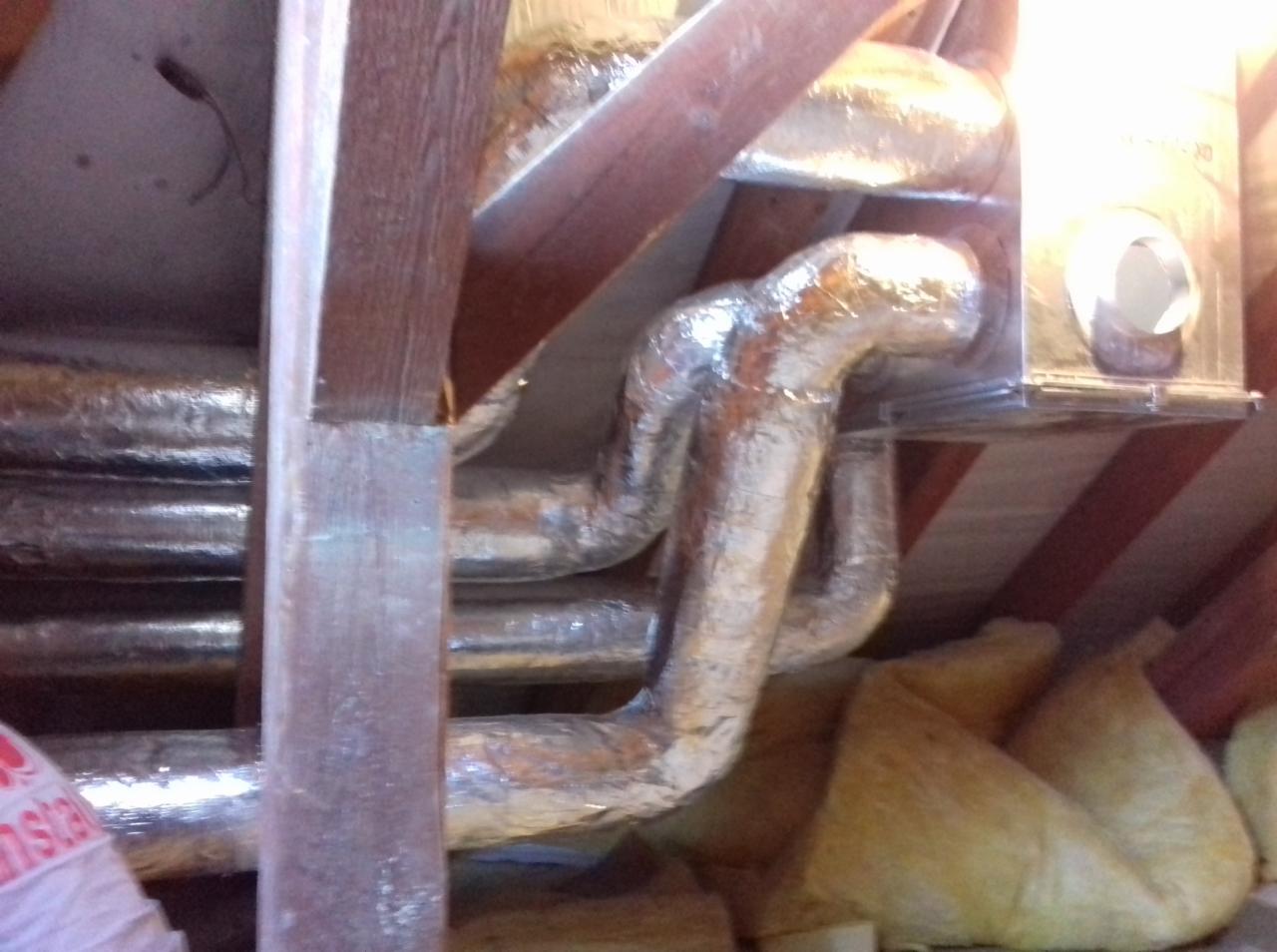 Srebrne, izolowane rury wentylacyjne poprowadzone pod drewnianym stropem, widoczna wełna mineralna.