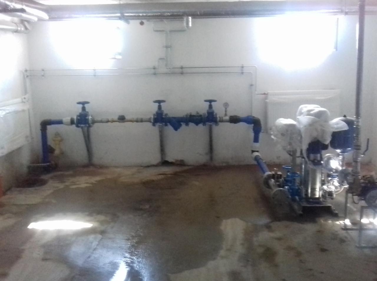 Piwnica z zainstalowanym systemem rur i zaworów, widoczne również pompy owinięte folią ochronną, na podłodze ślady wilgoci.