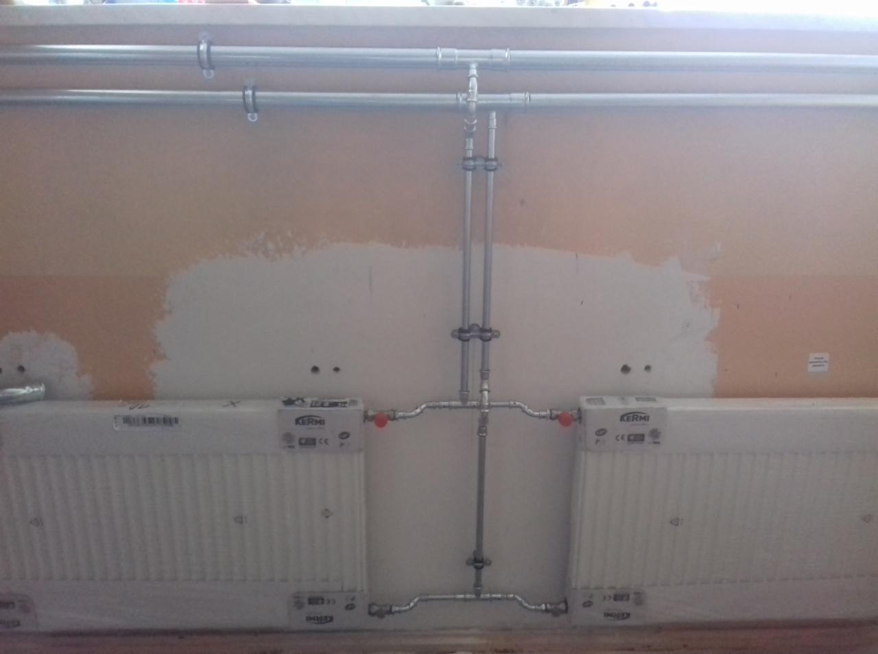 Dwa grzejniki marki Kermi zamontowane na ścianie z widocznymi rurami instalacji grzewczej, ściana częściowo pomalowana. Instalacja jeszcze nieukończona.