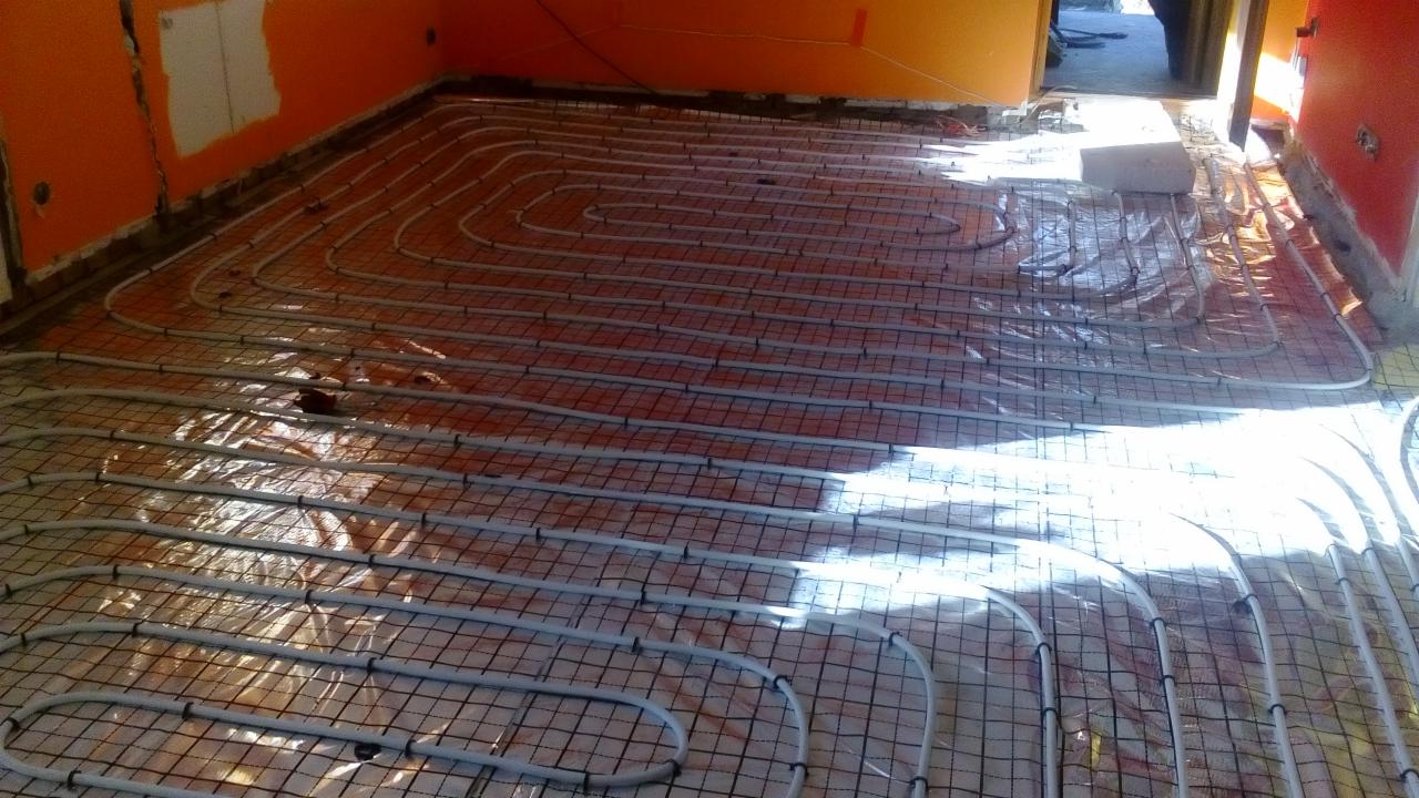 Instalacja ogrzewania podłogowego z rur rozprowadzonych na siatce w pomieszczeniu w trakcie remontu, pomarańczowe ściany w tle.