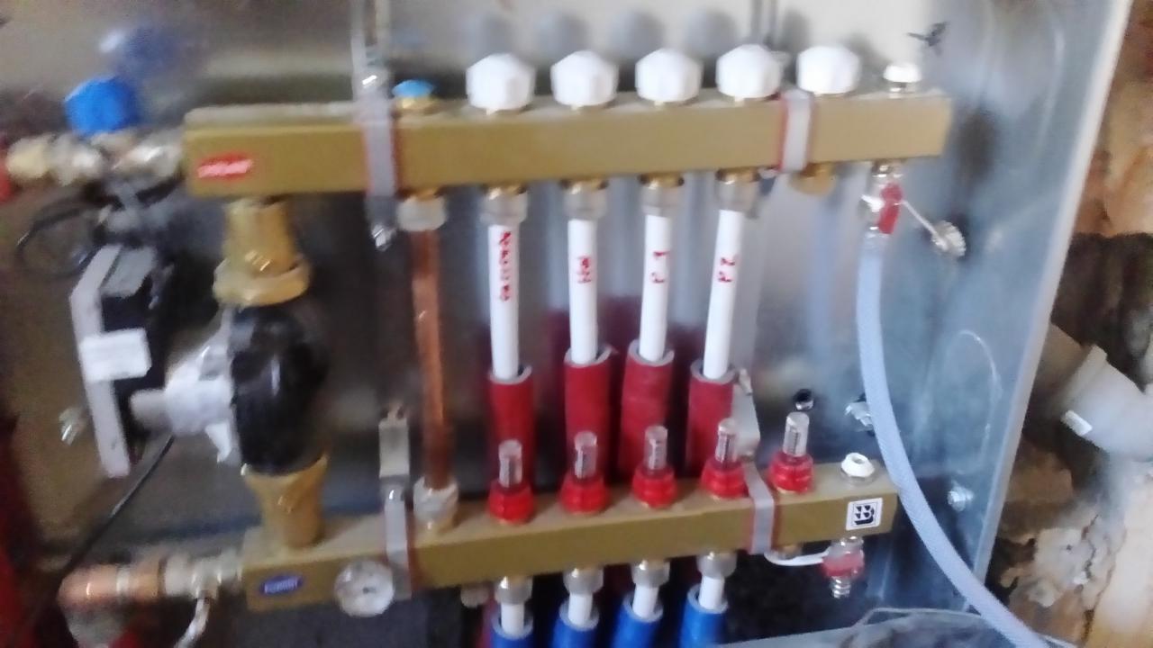 Rozdzielacz podłogowy z rurami oznaczonymi napisami, zamontowany na metalowej płycie, widoczne zawory i manometr.