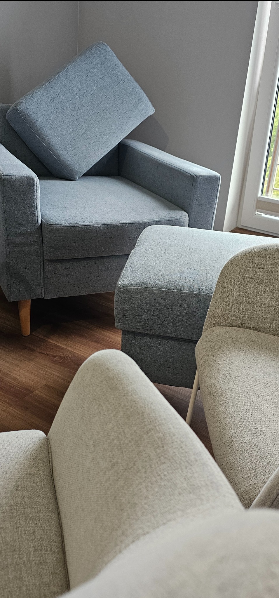 Szary fotel z poduszką i podnóżkiem, obok beżowe krzesło na drewnianej podłodze, widok z góry na fragment oparcia drugiego krzesła.