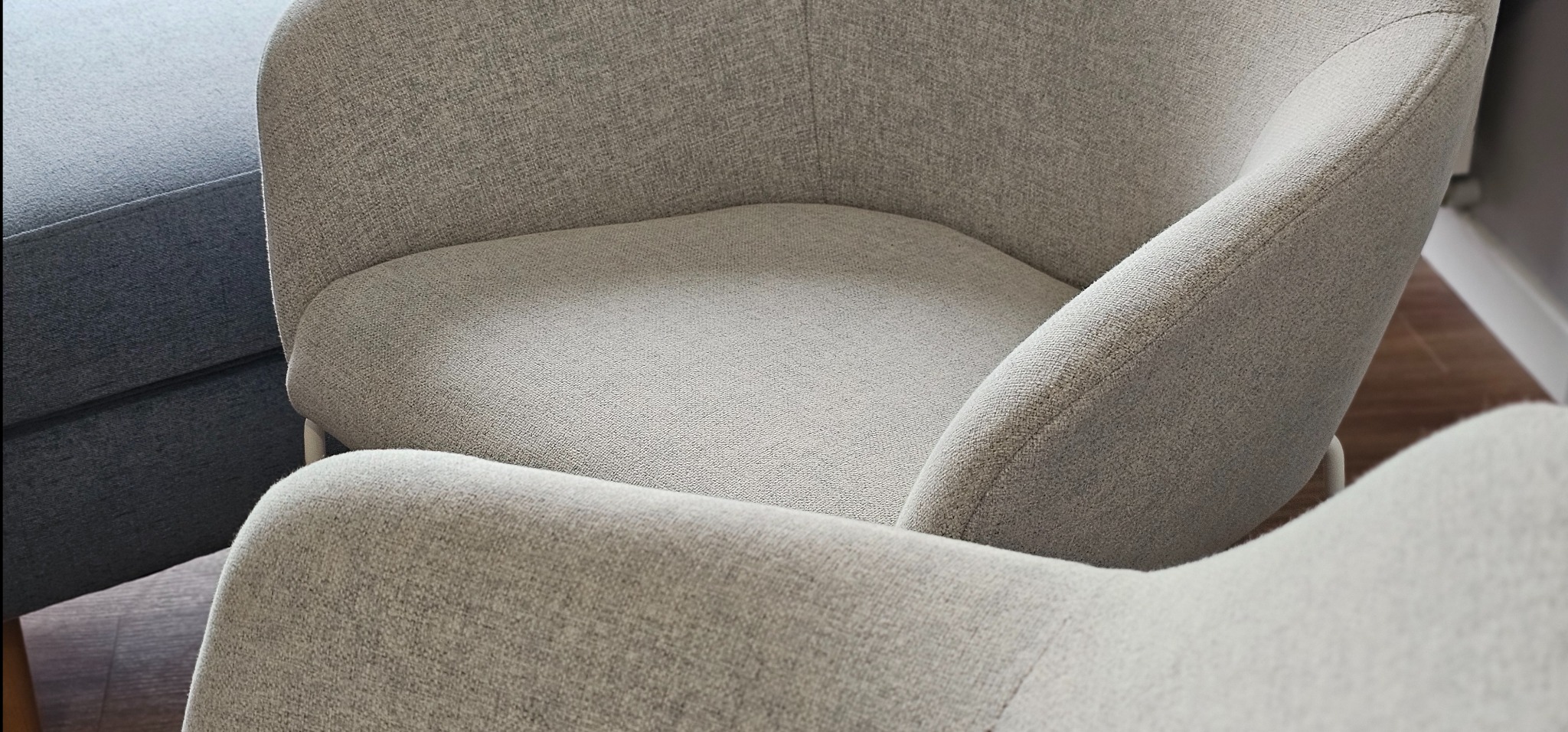 Fragment jasnoszarego fotela tapicerowanego z widoczną fakturą materiału, obok ciemnoszary puf, na drewnianej podłodze.