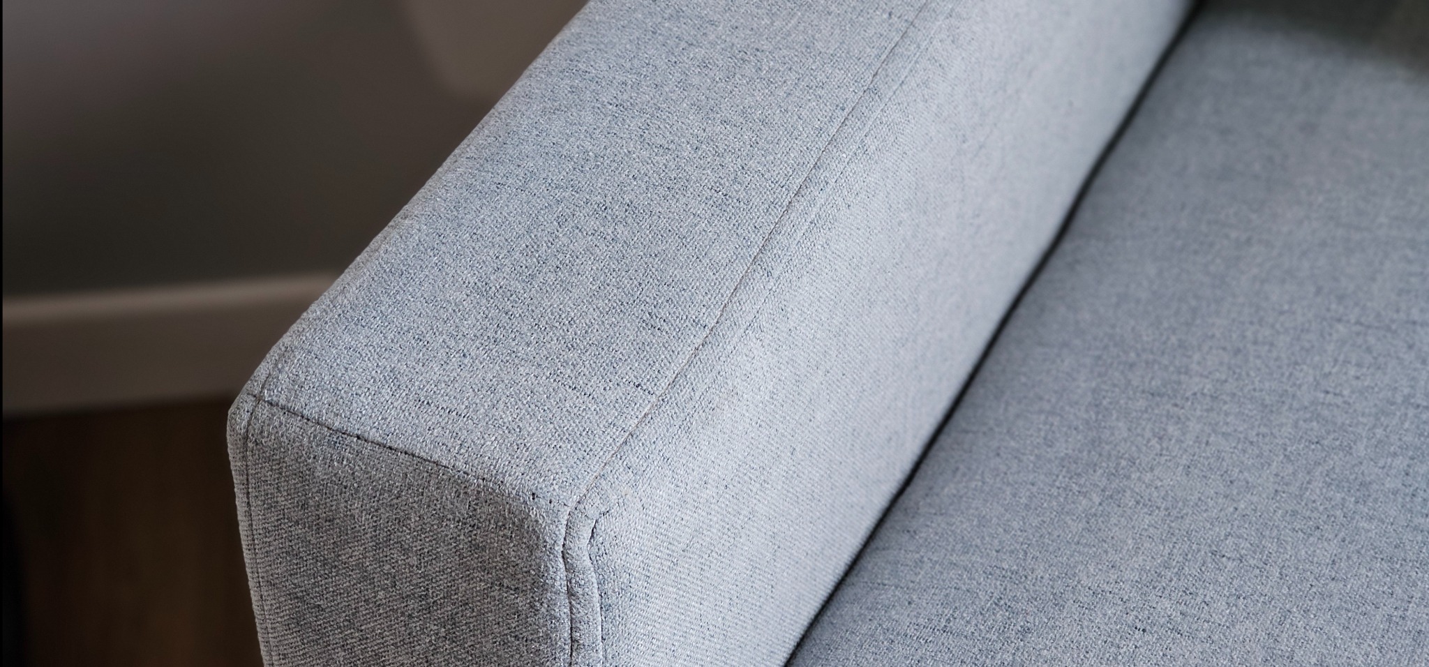Fragment jasnoniebieskiego podłokietnika sofy z widoczną fakturą tkaniny i szwem wykończeniowym, w tle ściana w neutralnym odcieniu szarości.