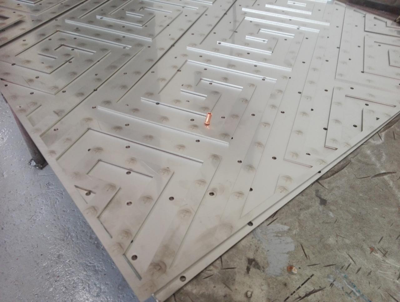 Stos arkuszy z wyciętymi laserowo labiryntami i otworami, widoczne ślady po obróbce, fragment miedzianego elementu na jednym z arkuszy.