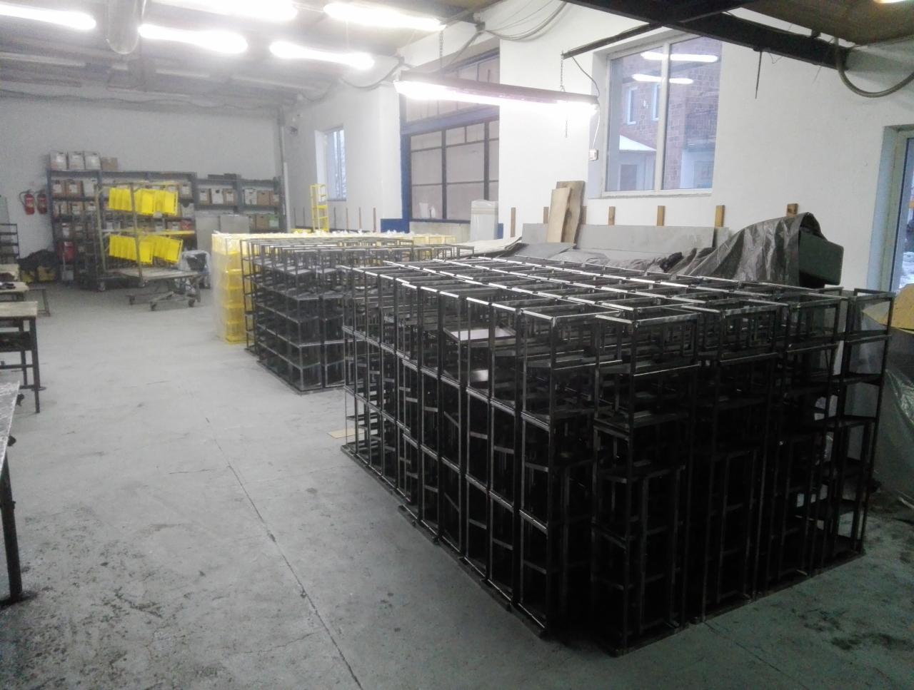 Stosy spawanych, metalowych konstrukcji szkieletowych w hali magazynowej, widoczne regały z żółtymi elementami w tle.