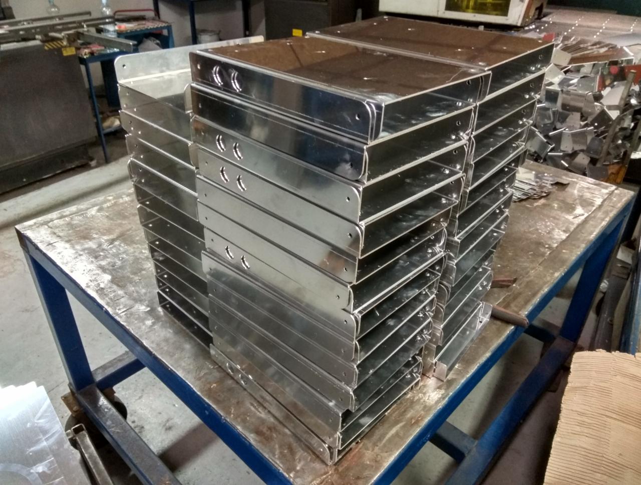 Stos precyzyjnie giętych, metalowych obudów z otworami, ułożonych na stalowym stole warsztatowym w hali produkcyjnej.