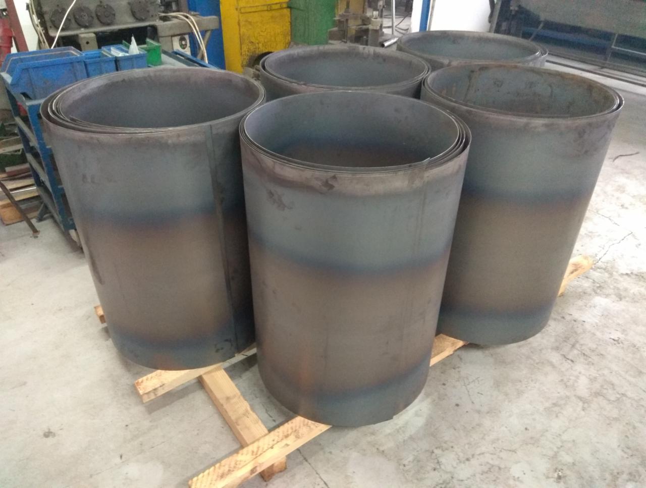 Walcowane blachy zwinięte w cylindryczne formy, ułożone na drewnianych legarach w hali produkcyjnej. Widoczne ślady spawania na krawędziach.