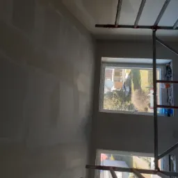 Ściana po nałożeniu gładzi, widoczne nierówności przed szlifowaniem, z fragmentem rusztowania i widokiem z okna na zewnątrz.