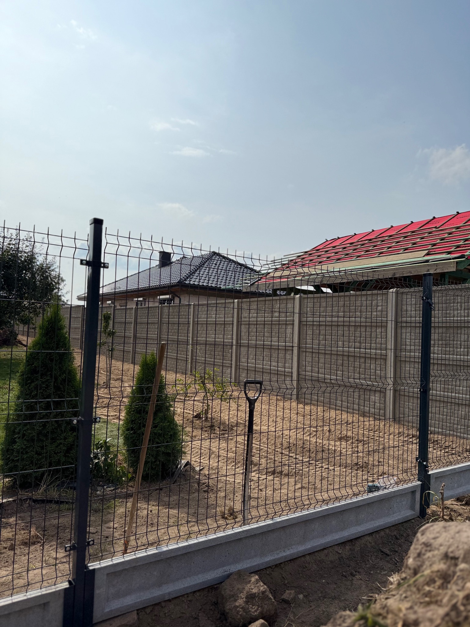 Nowoczesne ogrodzenie panelowe z podmurówką betonową na tle domów w budowie i nieba. W tle widać szpadel wbity w ziemię.