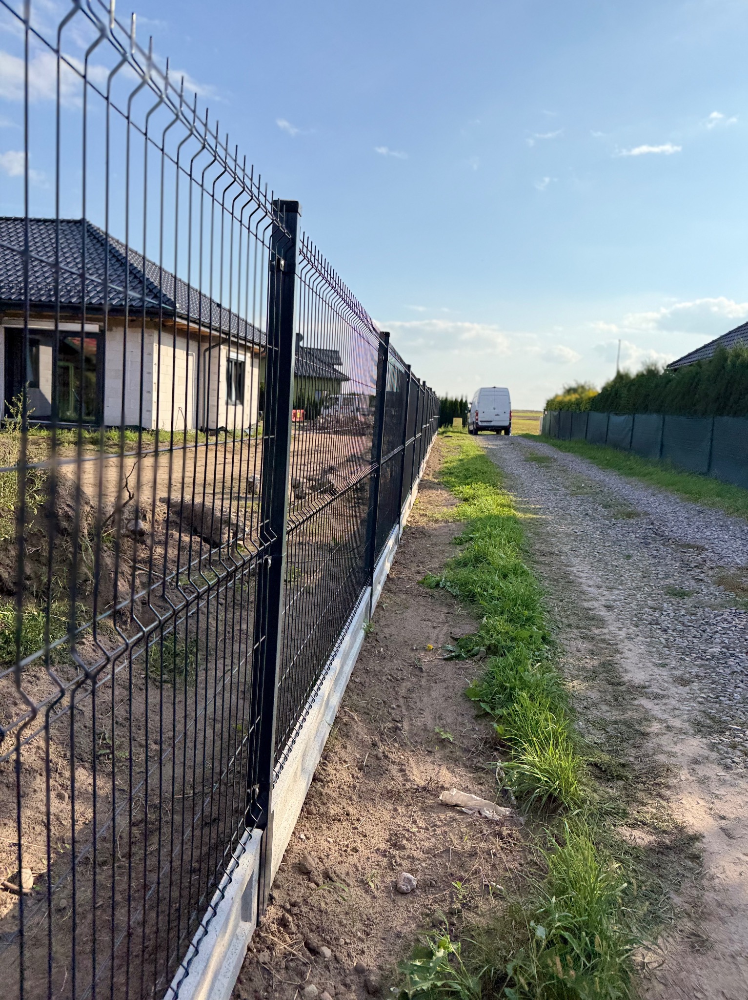 Ciemny panel ogrodzeniowy z podmurówką betonową, widok z boku na tle domu i drogi gruntowej z białym busem w oddali, w słoneczny dzień.