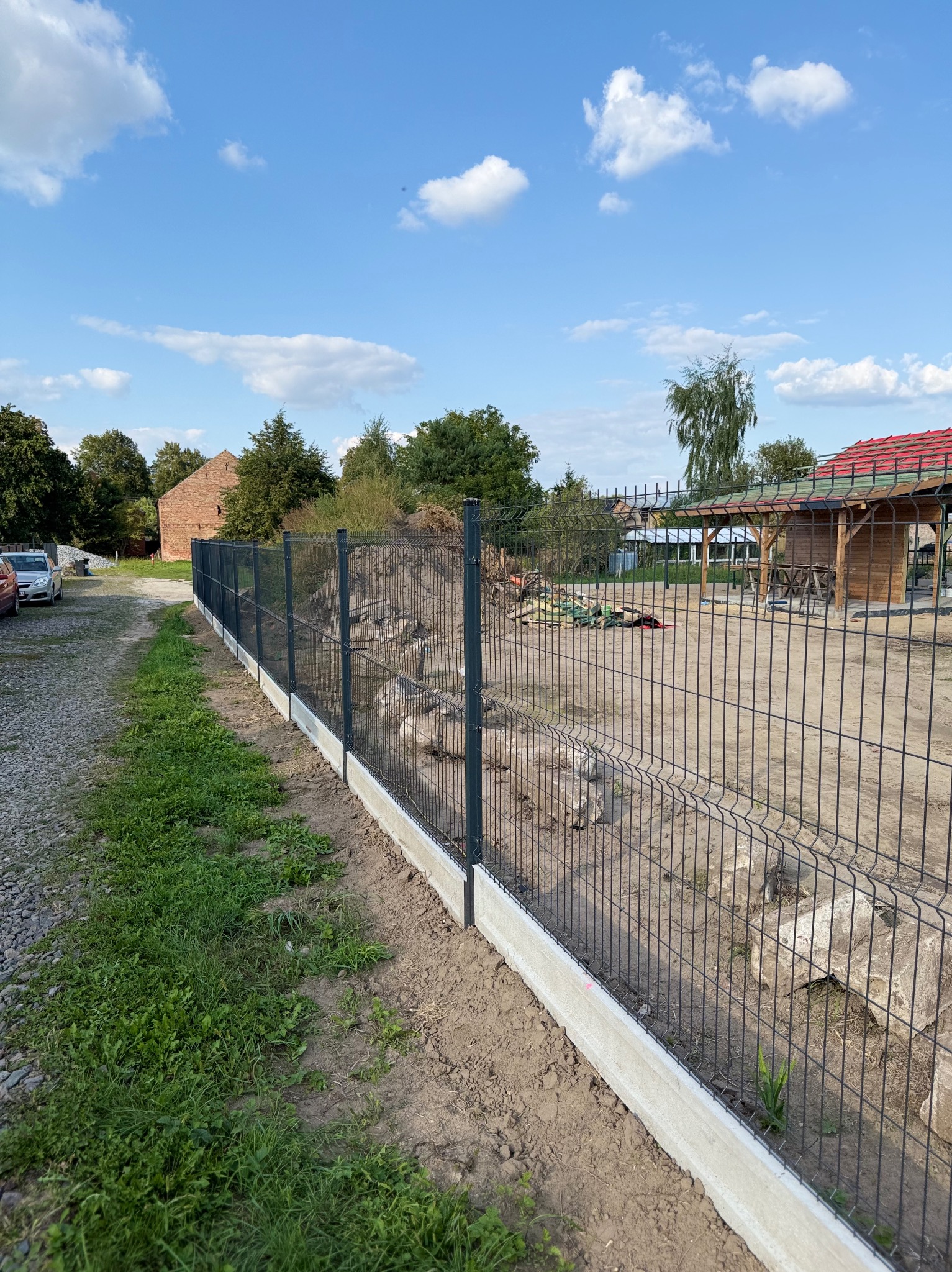 Nowoczesne ogrodzenie panelowe z podmurówką betonową na działce. Widok na zielony teren i zabudowania w tle, błękitne niebo z chmurami.