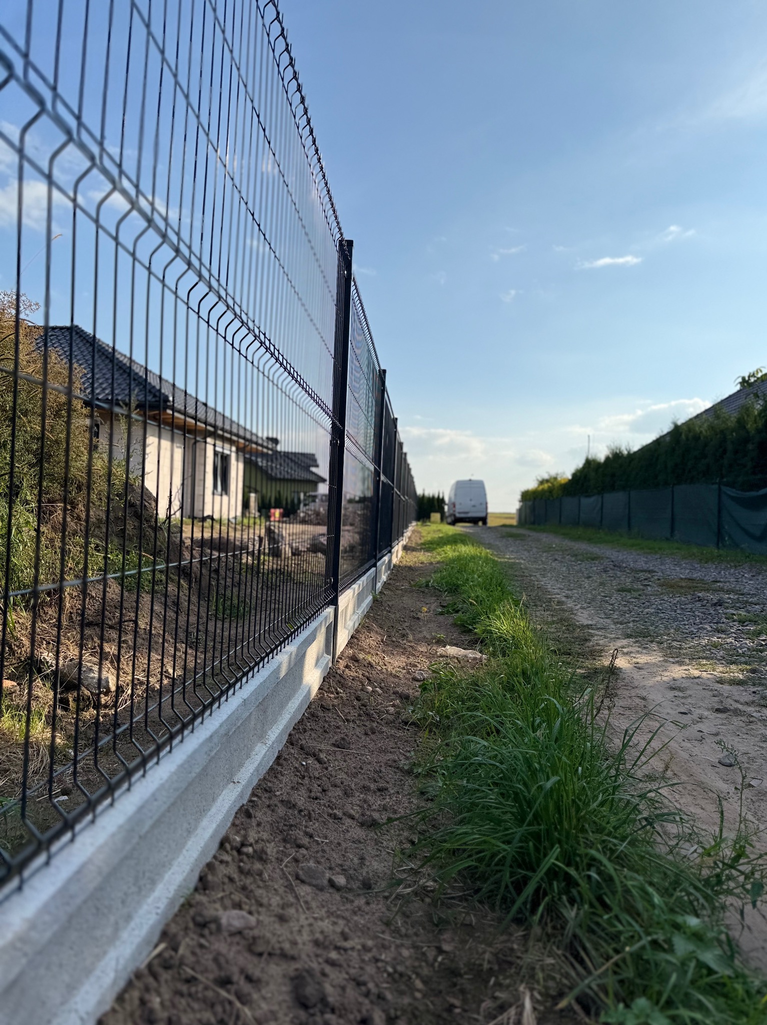 Nowoczesne ogrodzenie panelowe z podmurówką betonową, widok z perspektywy gruntu na tle domu i błękitnego nieba z białym busem w oddali.