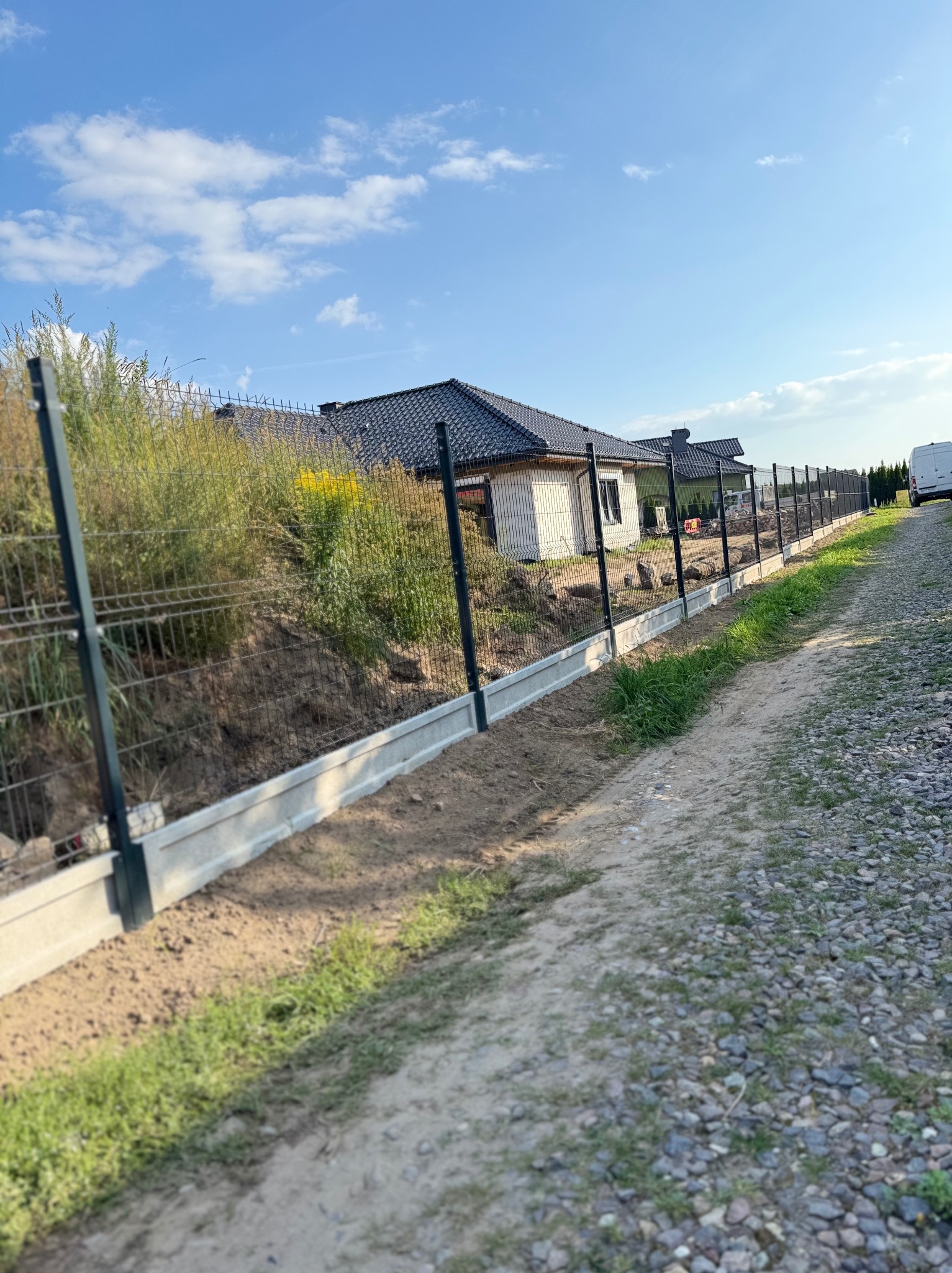 Montaż ogrodzenia panelowego z podmurówką betonową wzdłuż posesji. Widoczne panele i słupki w kolorze grafitowym, a w tle dom z ciemnym dachem i błękitne niebo.