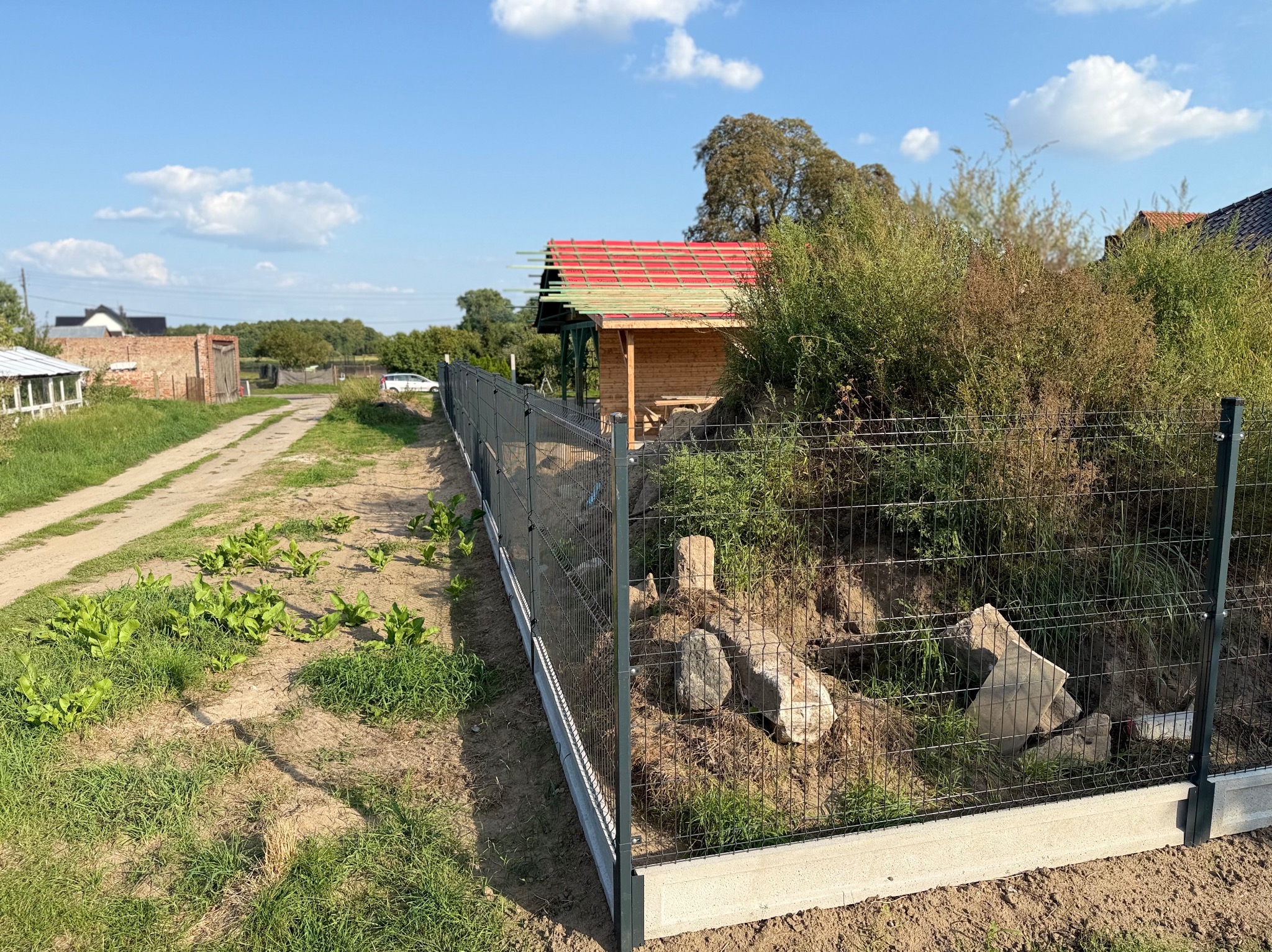 Montaż ogrodzenia panelowego na nierównym terenie, widoczne fundamenty betonowe, zielona trawa i budynek w tle. Ujęcie w słoneczny dzień.