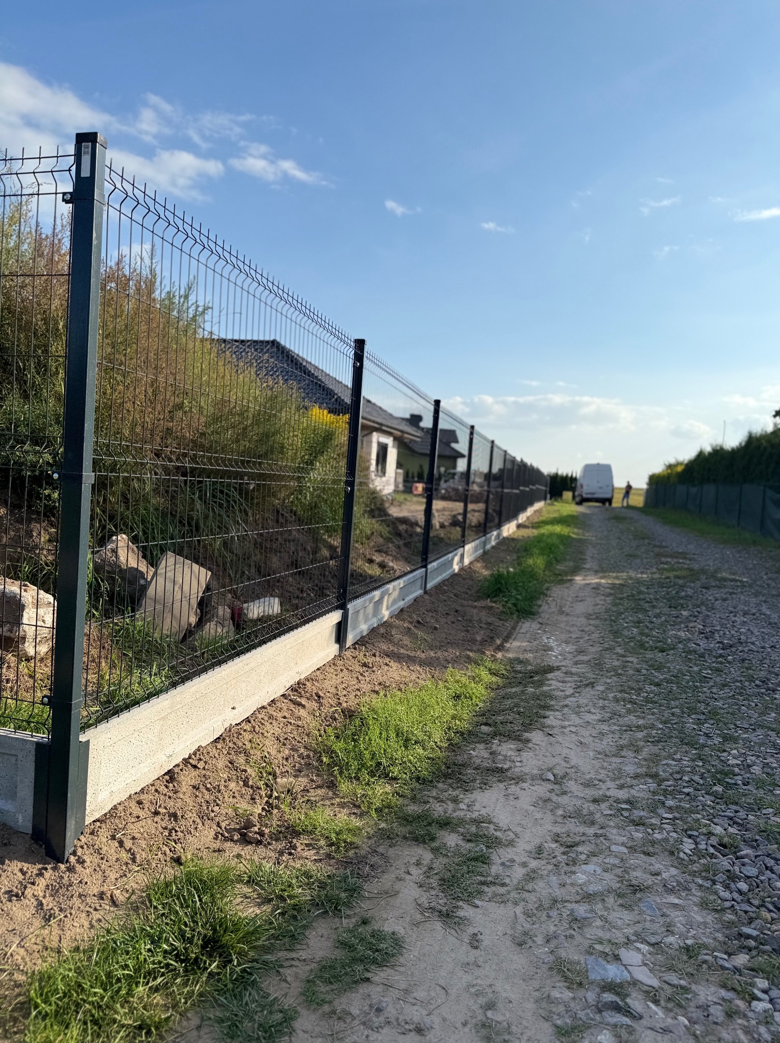 Długi, szary panelowy płot z metalową siatką, ciągnący się wzdłuż polnej drogi. W tle dom i błękitne niebo. Widoczna perspektywa zbieżna.