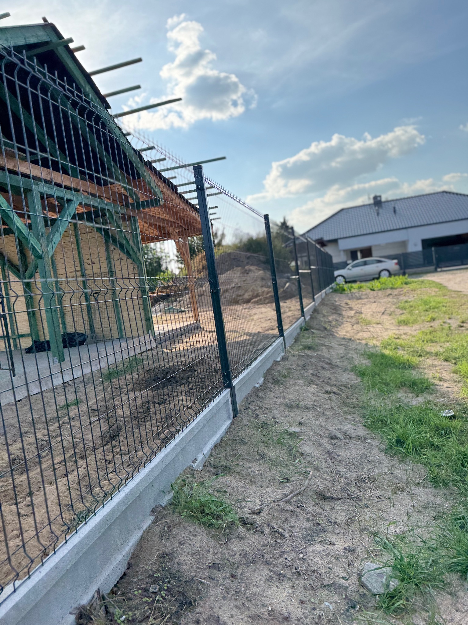Nowoczesne ogrodzenie panelowe na betonowej podmurówce, widok z perspektywy gruntu, z domem w tle i elementami szkieletu budynku w budowie.