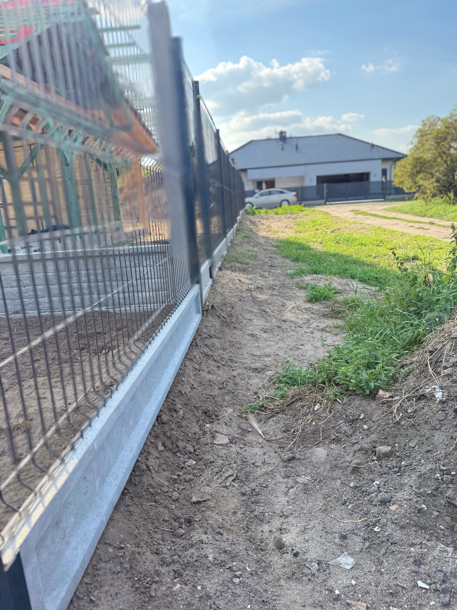 Nowoczesne ogrodzenie panelowe z podmurówką betonową, widok z bliska na grunt i elementy mocujące. W tle dom jednorodzinny i błękitne niebo.