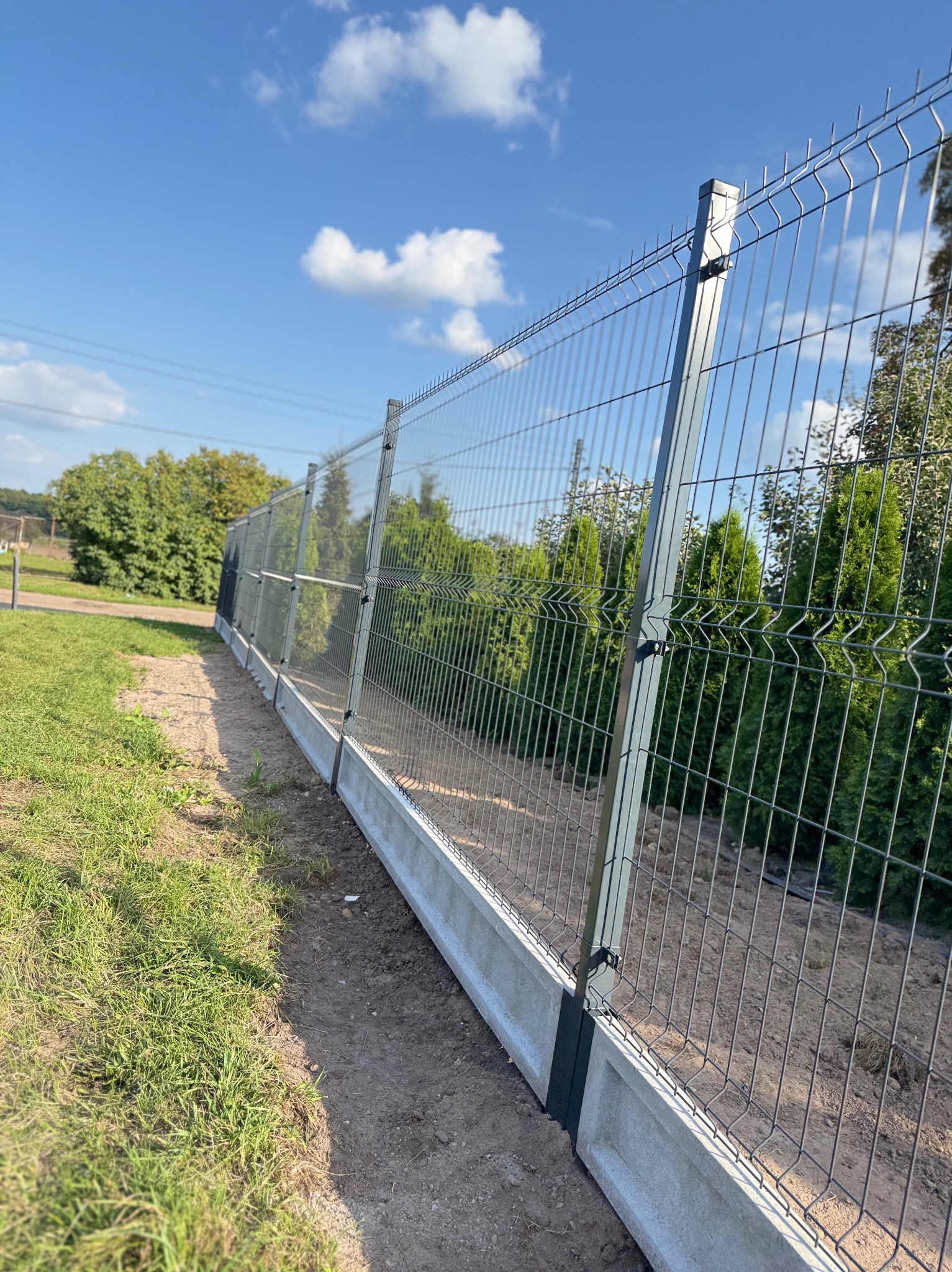 Nowoczesne ogrodzenie panelowe z podmurówką betonową, widok z boku na tle zieleni i błękitnego nieba. Metalowa siatka ogrodzeniowa w szarym kolorze.