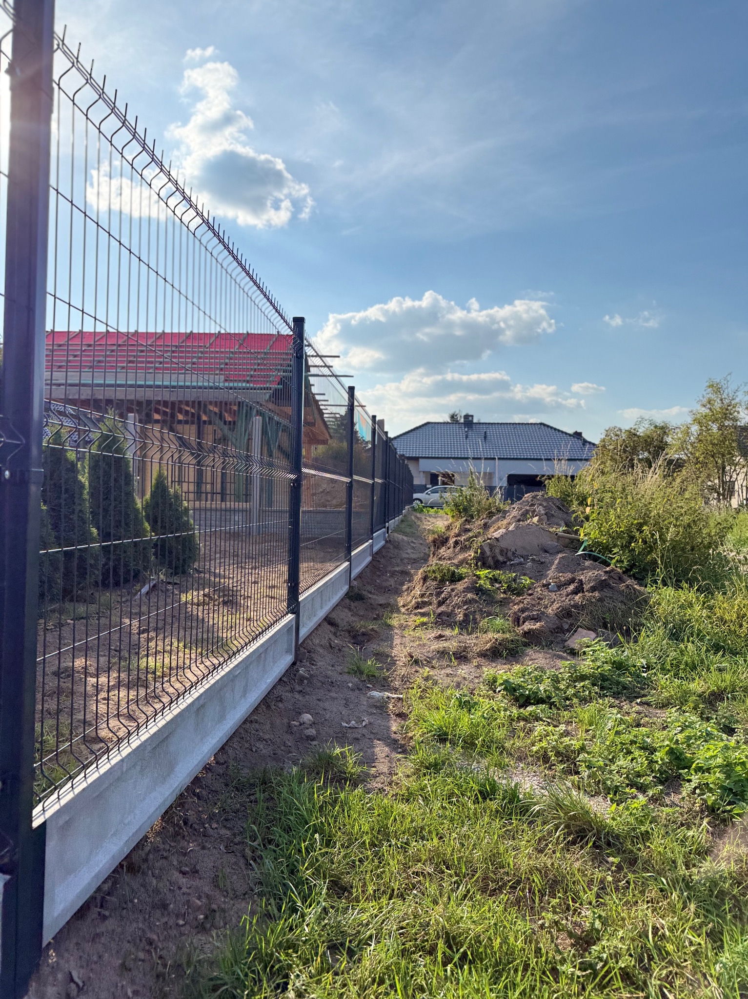 Nowoczesne ogrodzenie panelowe z podmurówką betonową, widok z perspektywy gruntu, błękitne niebo w tle. Słoneczny dzień, świeżo posadzona zieleń.