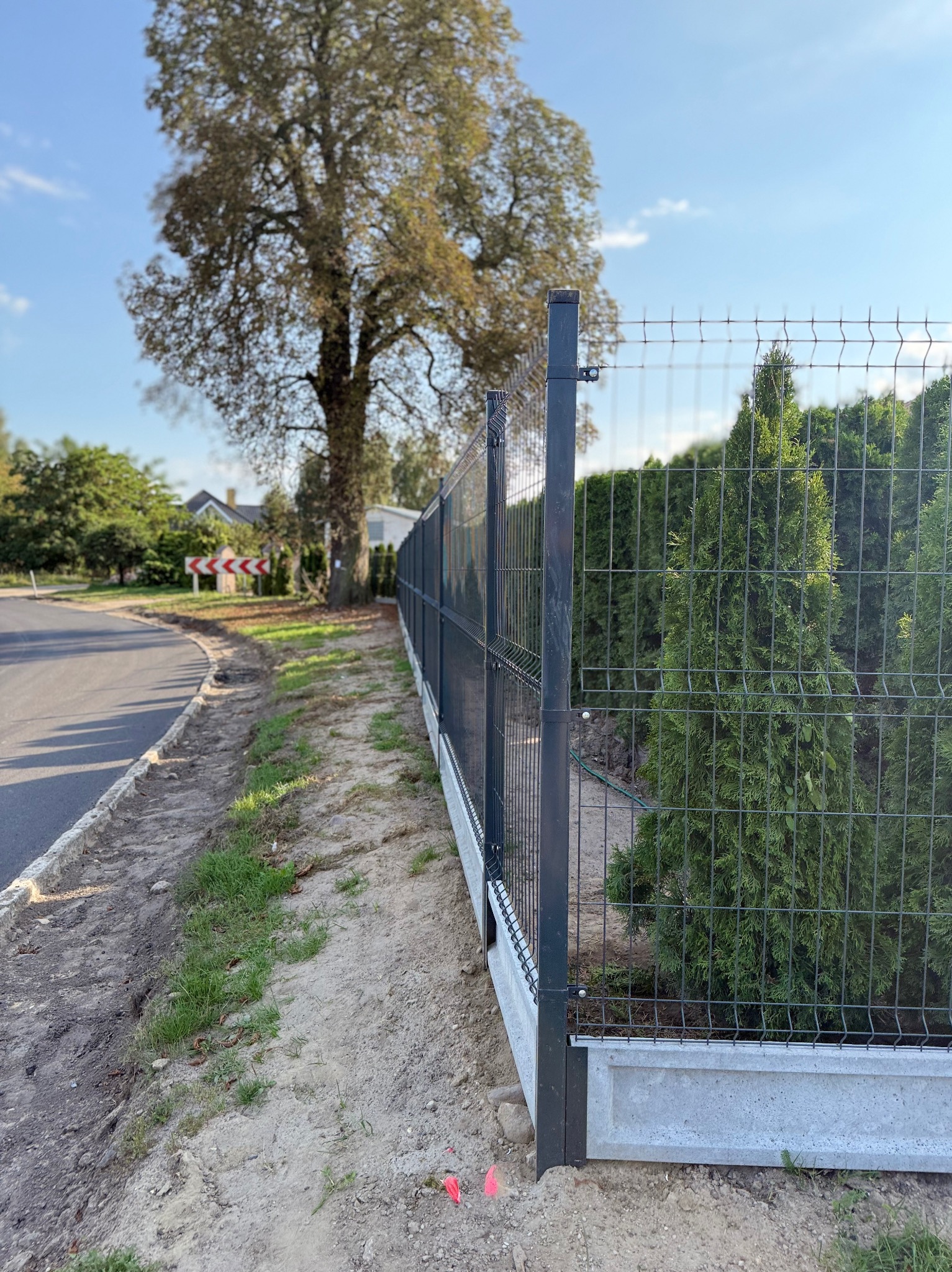 Nowoczesne ogrodzenie panelowe z podmurówką betonową w Kobylance. Widok z boku, zieleń w tle. Ciemnoszary metal kontrastuje z jasną podmurówką i zielenią.