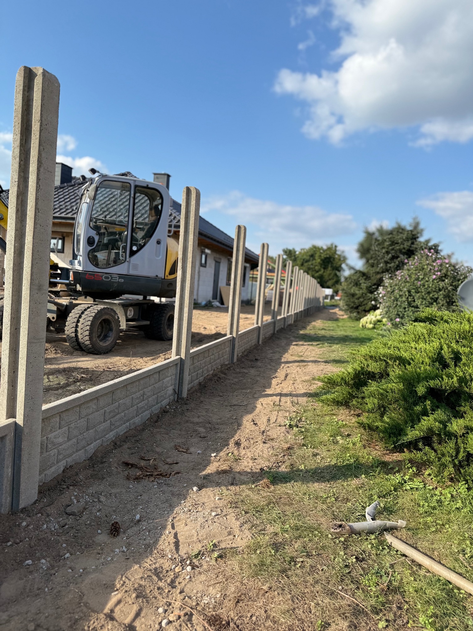 Montaż ogrodzenia panelowego z podmurówką i słupkami betonowymi. W tle minikoparka i dom jednorodzinny w słoneczny dzień.