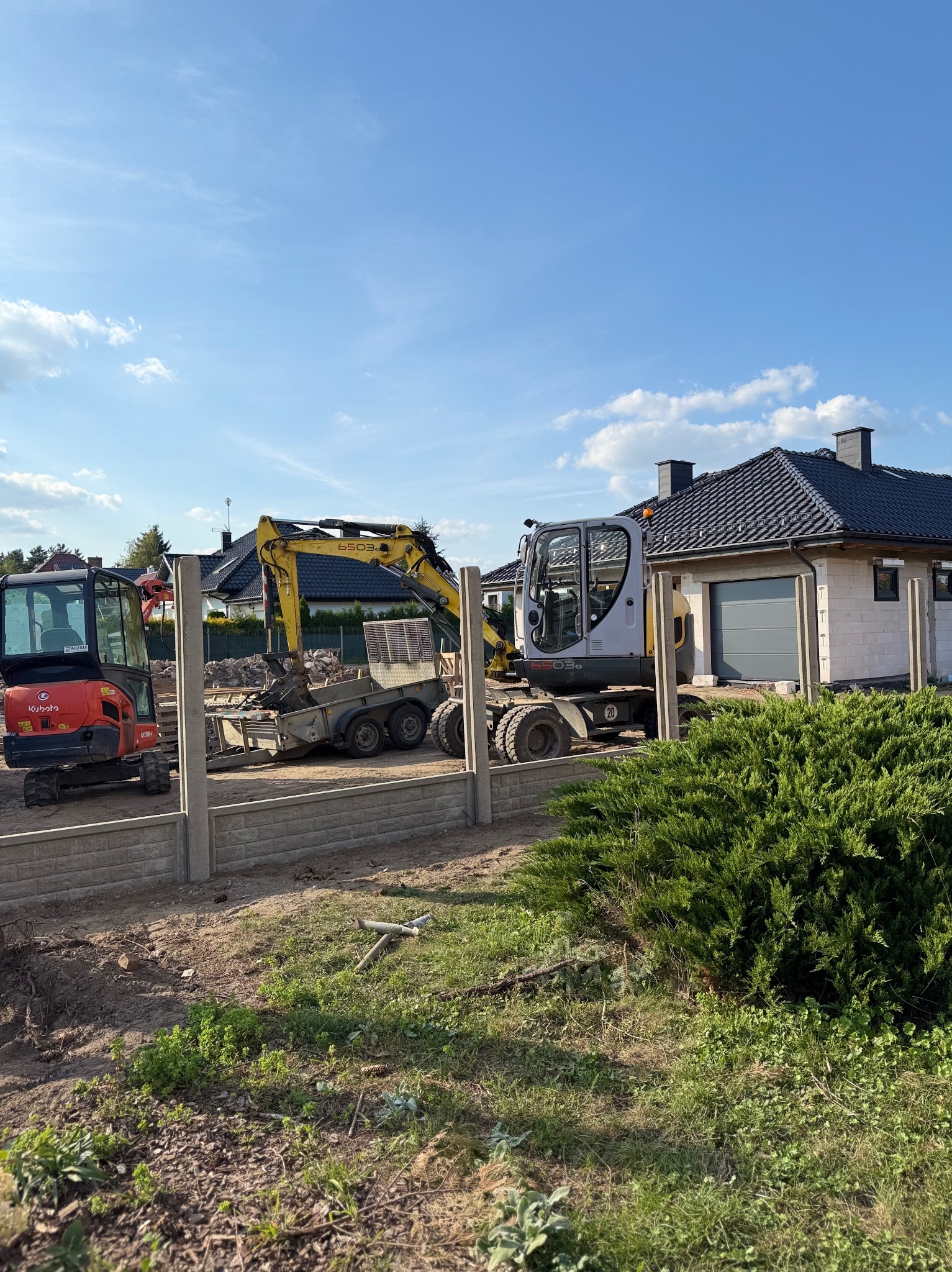 Montaż ogrodzenia panelowego z użyciem mini koparek na tle domu jednorodzinnego w słoneczny dzień. Widoczne słupki i częściowo zamontowane panele.