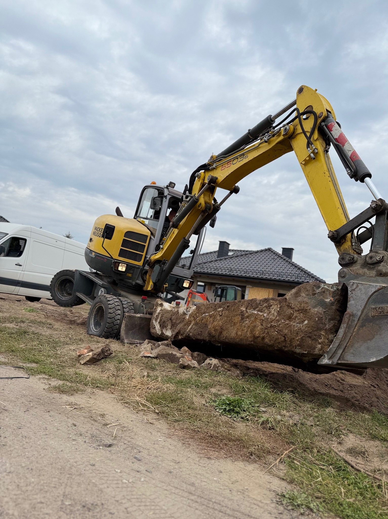 Żółta koparka Wacker Neuson 6503 wykopuje ziemię na tle domu i białego busa. Widok z dołu, pochmurne niebo.