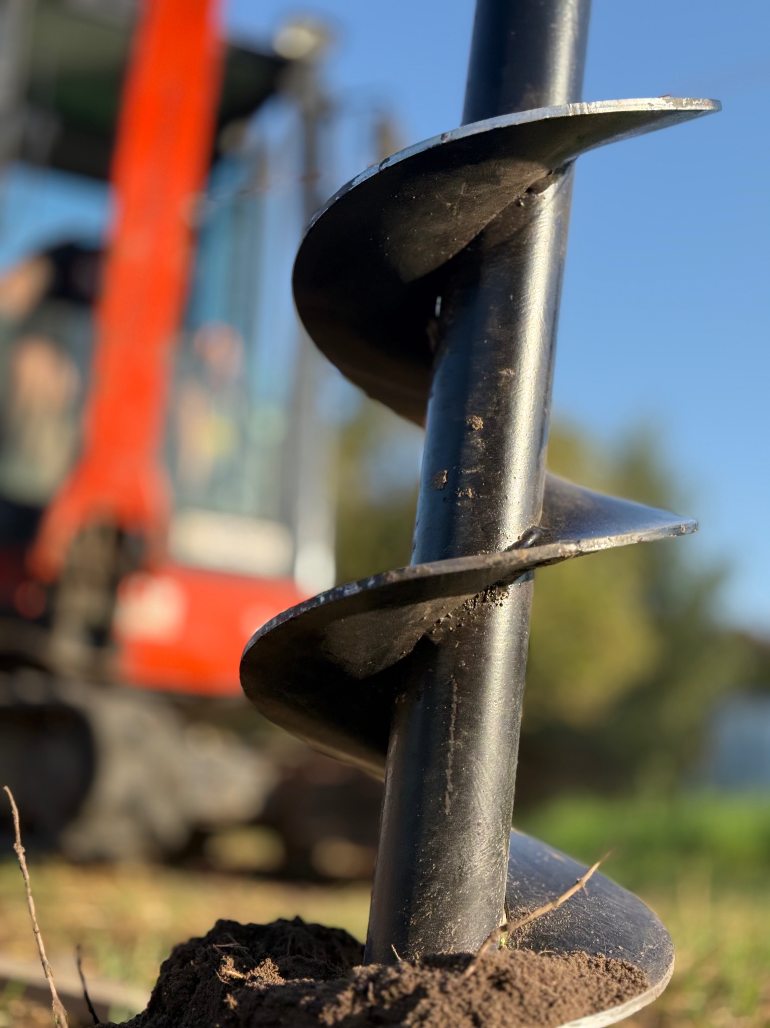 Wiertnica do gruntu z bliska, z pomarańczową koparką w tle i błękitnym niebem. Wiertło wbite w ziemię, gotowe do pracy przy instalacji ogrodzenia.