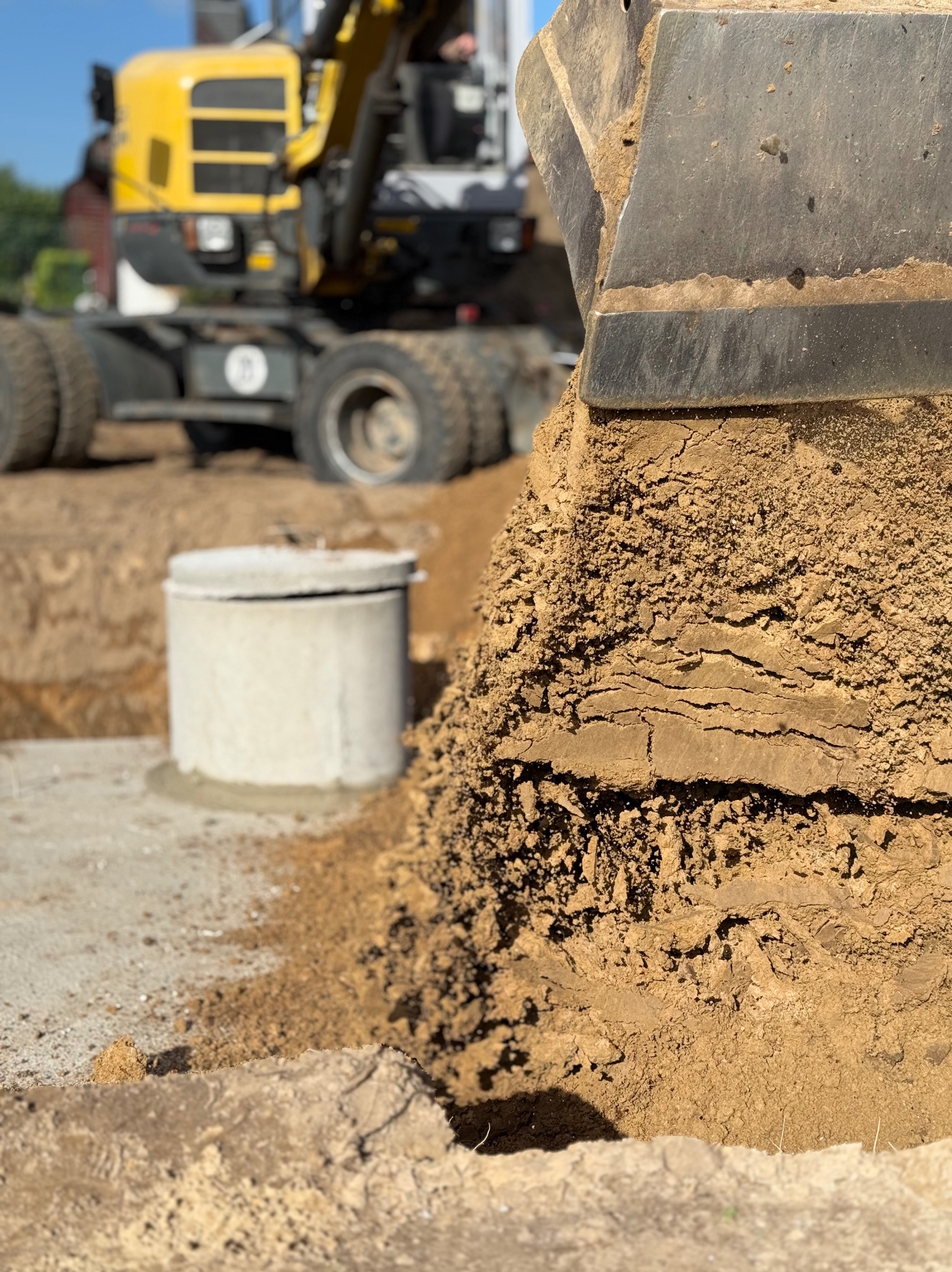 Żółta koparka z łyżką pełną ziemi, w tle betonowy krąg studzienny i wykop. Prace ziemne przyłącza wod-kan.