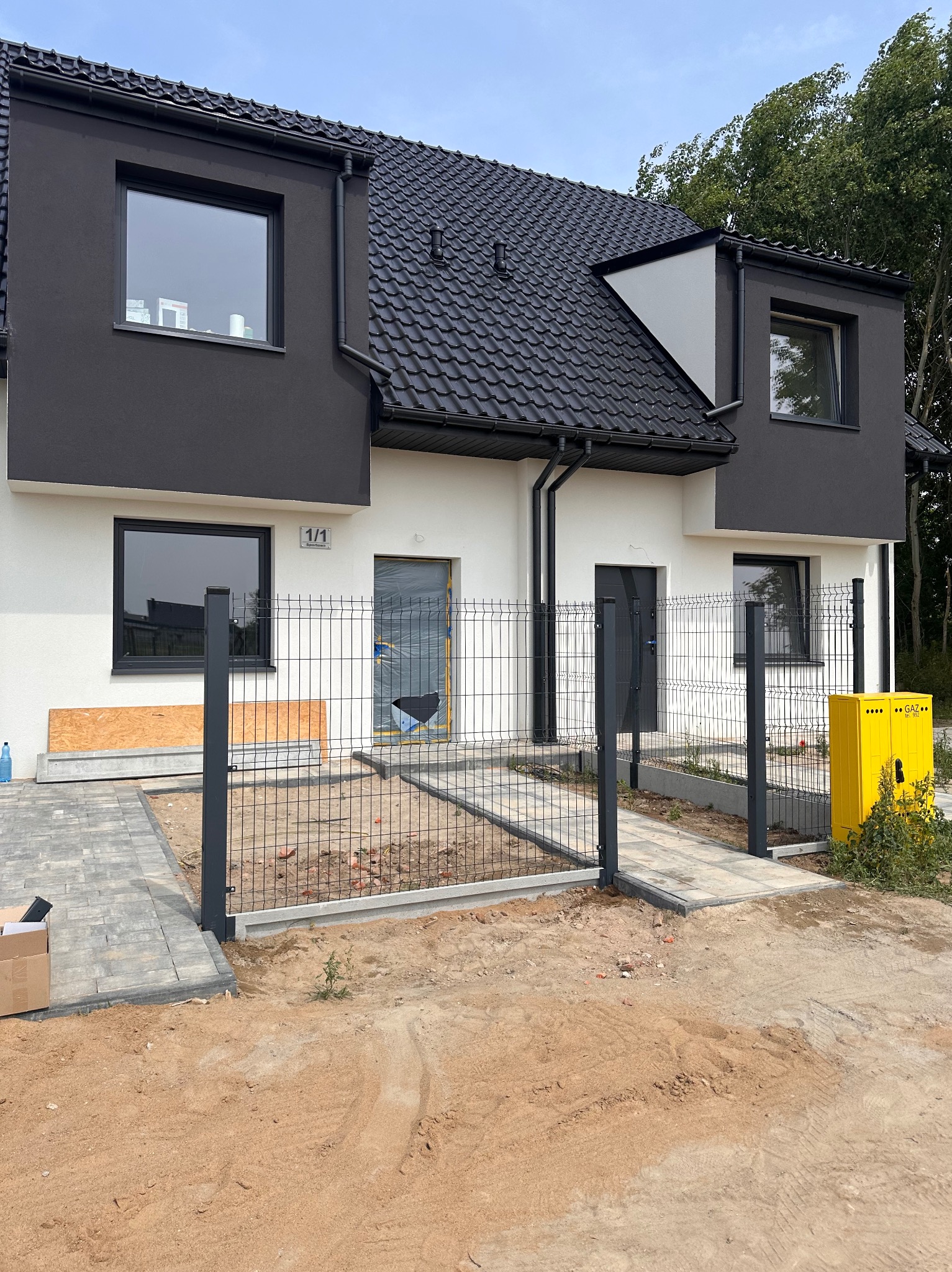 Nowoczesny bliźniak z czarną stolarką i szarym ogrodzeniem panelowym. Żółta skrzynka gazowa przy wejściu. Kostka brukowa i teren w trakcie prac ziemnych.