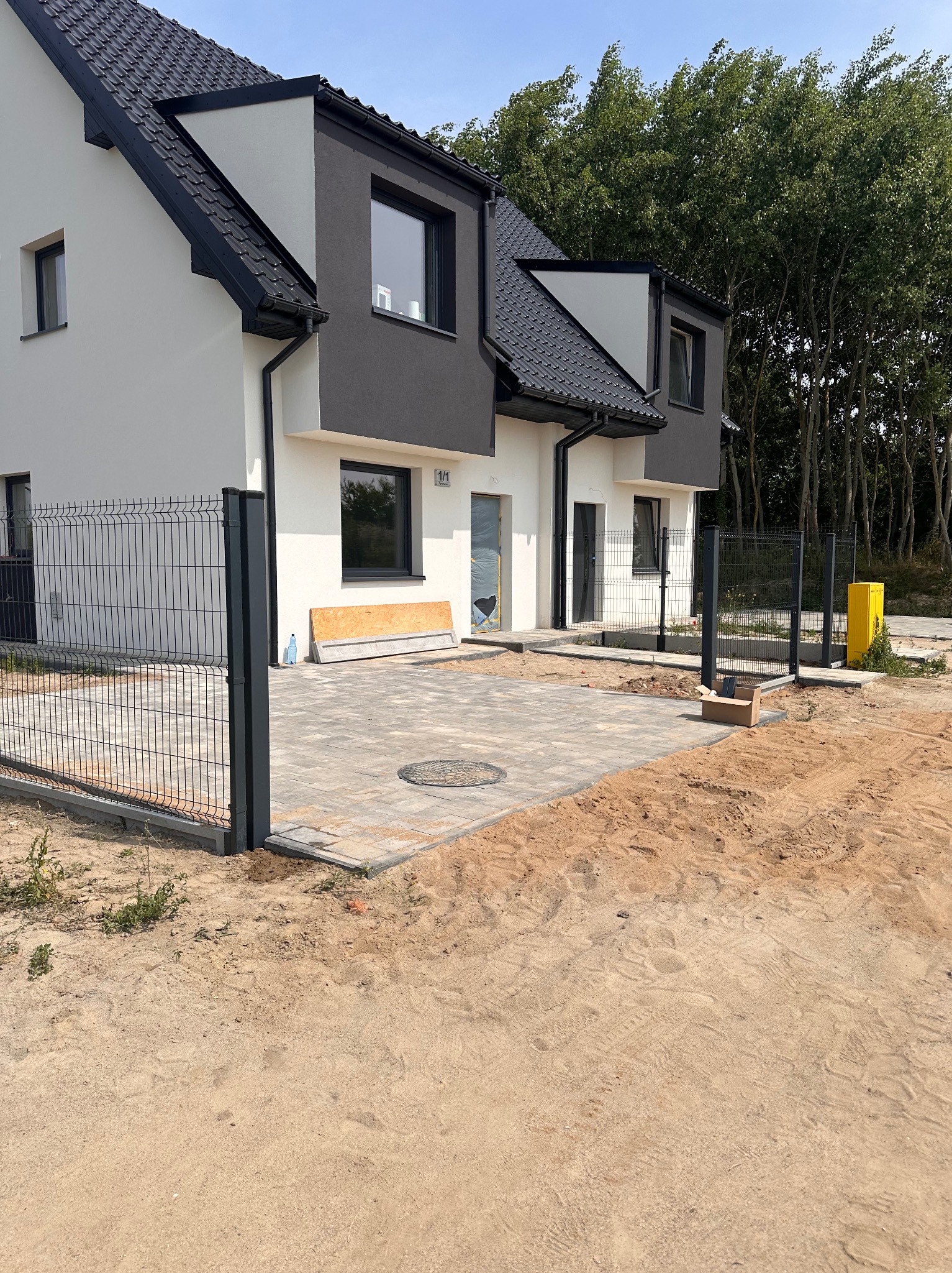 Nowoczesny dom szeregowy z grafitową elewacją, ogrodzeniem panelowym i wybrukowanym podjazdem z szarej kostki. Teren wokół w trakcie prac wykończeniowych.