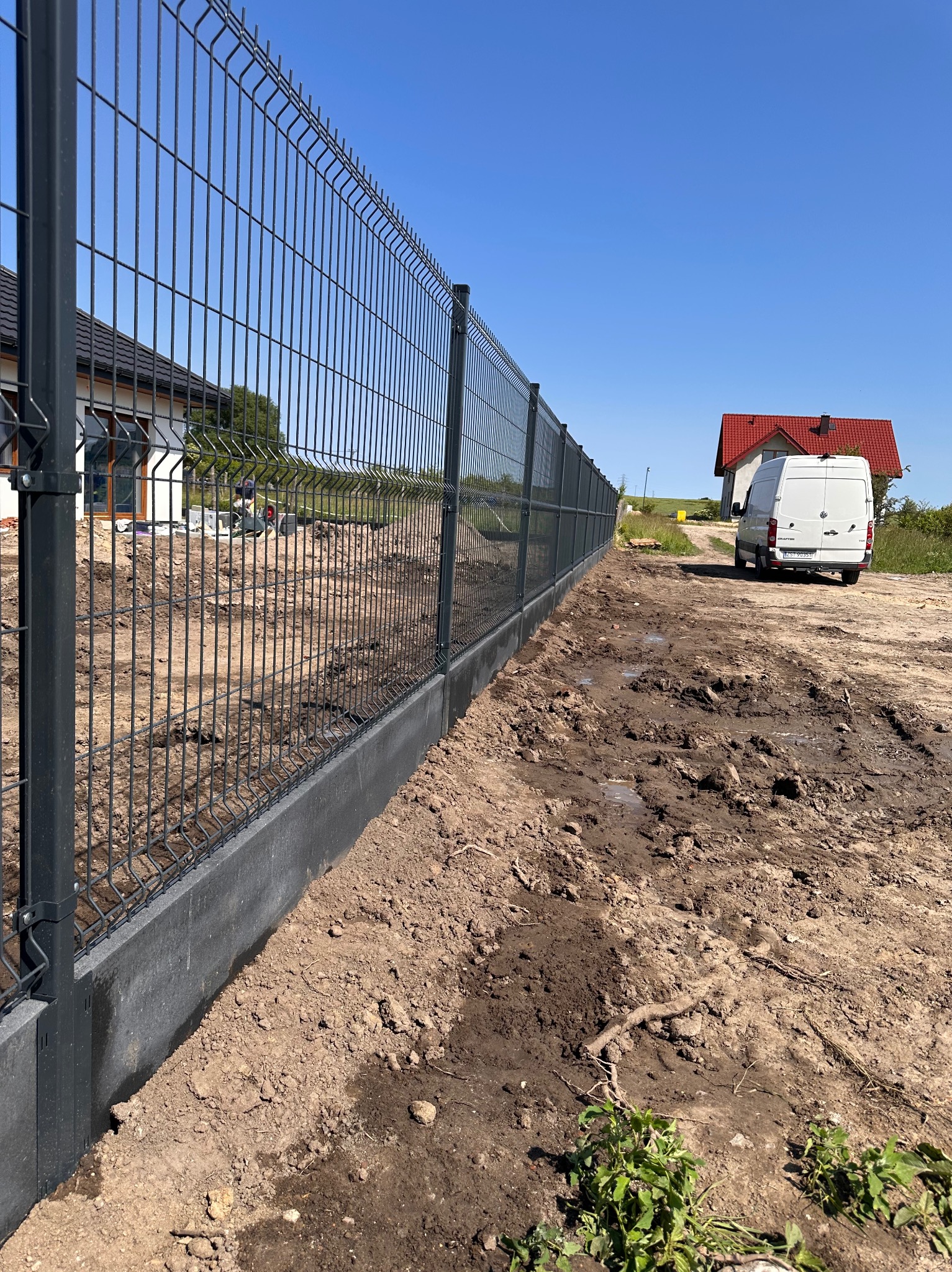 Nowoczesne ogrodzenie panelowe z podmurówką, widok z perspektywy gruntu, z białym busem w tle i fragmentem domu z czerwonym dachem. Słoneczny dzień.