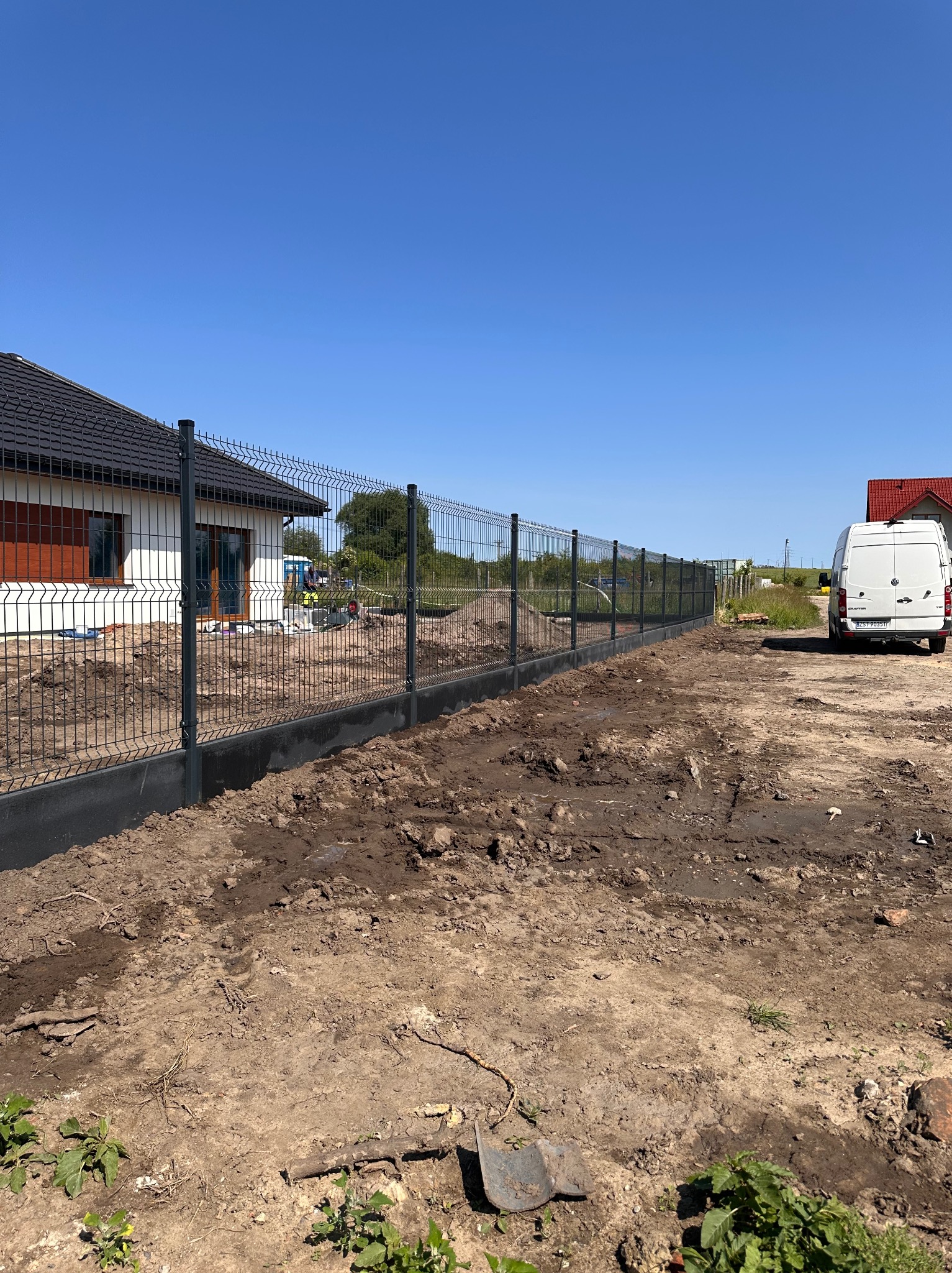 Nowoczesne ogrodzenie panelowe z podmurówką w kolorze grafitowym, otoczone świeżo przekopaną ziemią. W tle dom jednorodzinny i biały bus.