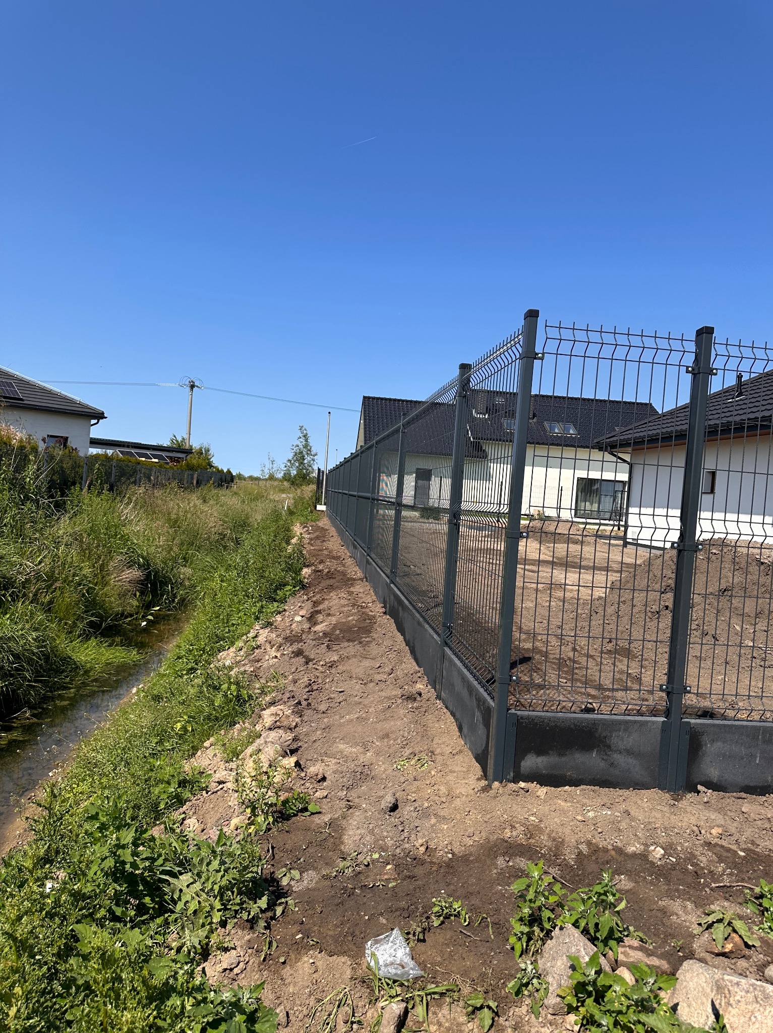 Nowe, szare ogrodzenie panelowe z podmurówką betonową, oddzielające posesję od cieku wodnego. W tle widoczne domy jednorodzinne pod błękitnym niebem.