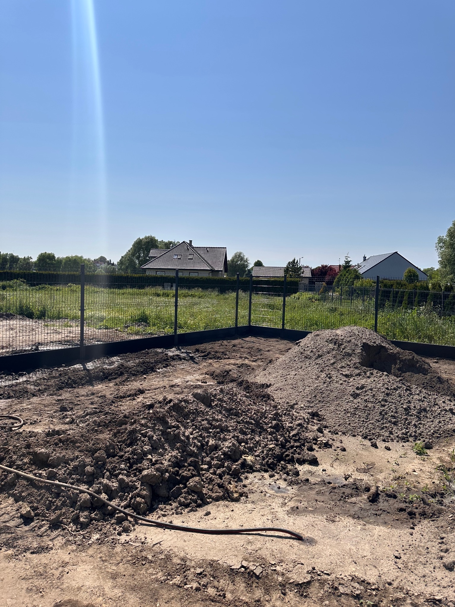 Montaż ogrodzenia panelowego na działce: widoczne słupki, podmurówka, ziemia i hałdy piasku. W tle domy i zieleń pod błękitnym niebem.