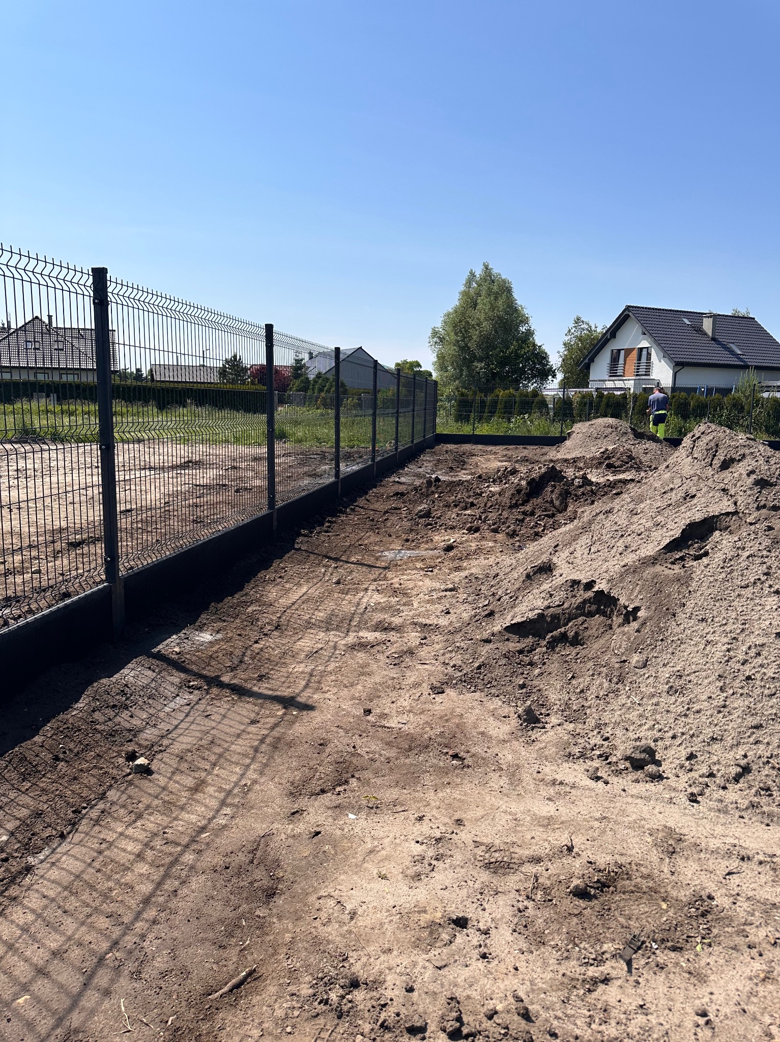 Montaż ogrodzenia panelowego: widok na wkopane słupki i częściowo zamontowane panele, hałdy ziemi wzdłuż linii ogrodzenia, dom w tle.