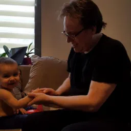 Mężczyzna w okularach delikatnie trzyma dłonie uśmiechniętego dziecka siedzącego na kanapie w salonie, w tle okno z roletami i rośliny doniczkowe.