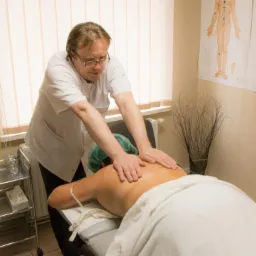 Mężczyzna w białym uniformie uciska plecy pacjentki leżącej na stole do masażu; widoczne plakaty anatomiczne na ścianie i wózek z akcesoriami.