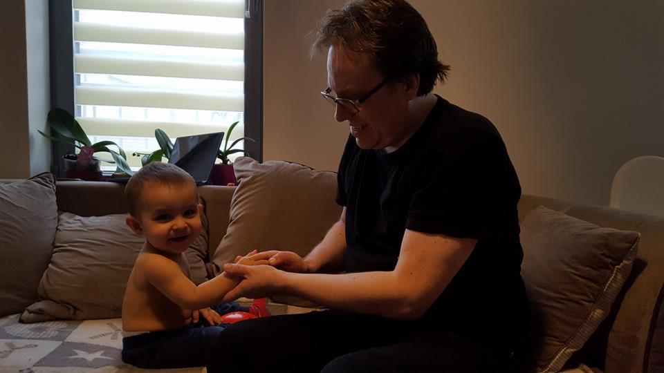 Mężczyzna w okularach delikatnie trzyma dłonie uśmiechniętego dziecka siedzącego na kanapie w salonie, w tle okno z roletami i rośliny doniczkowe.