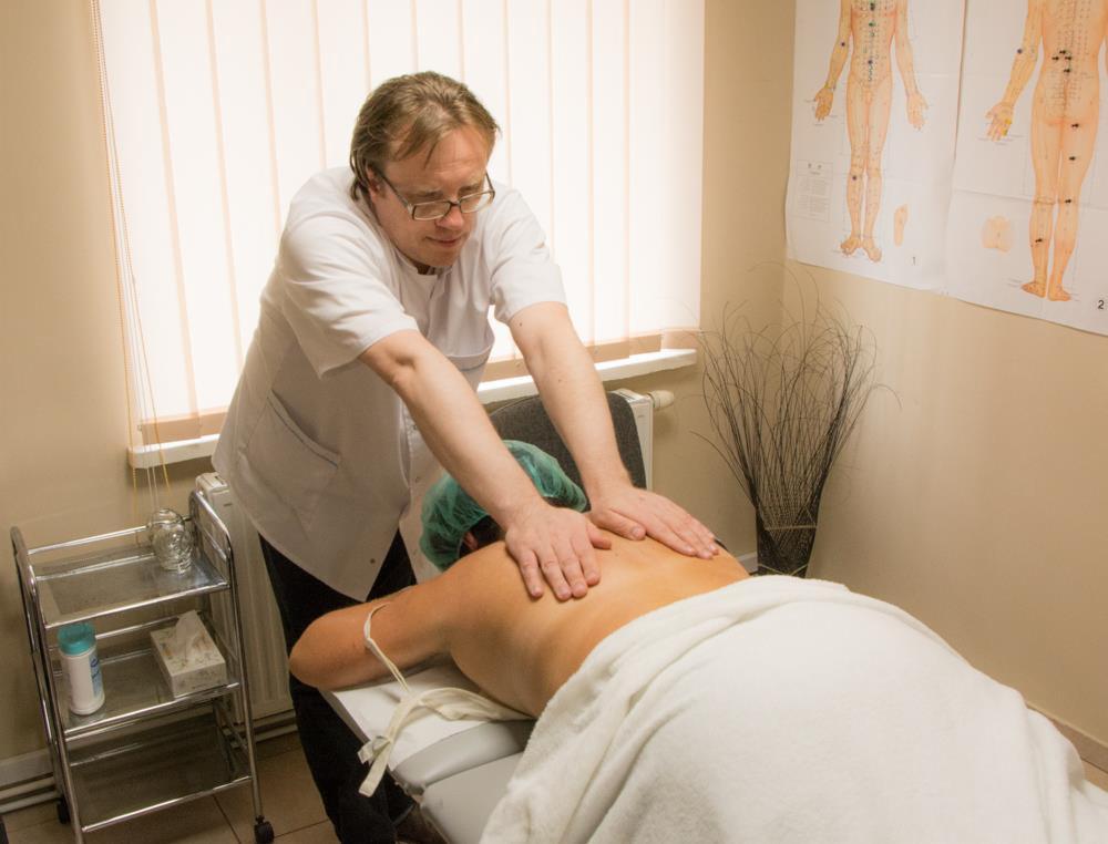 Mężczyzna w białym uniformie uciska plecy pacjentki leżącej na stole do masażu; widoczne plakaty anatomiczne na ścianie i wózek z akcesoriami.