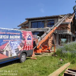 Montaż dachówki na nowym domu z użyciem podnośnika transportowego, widoczny samochód firmowy z reklamą pokryć dachowych i numerem telefonu, w tle kwitnące niebieskie kwiaty.