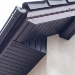 Fragment dachu z brązową blachą trapezową, orynnowaniem i podbitką, widok z dołu na tle jasnej ściany.