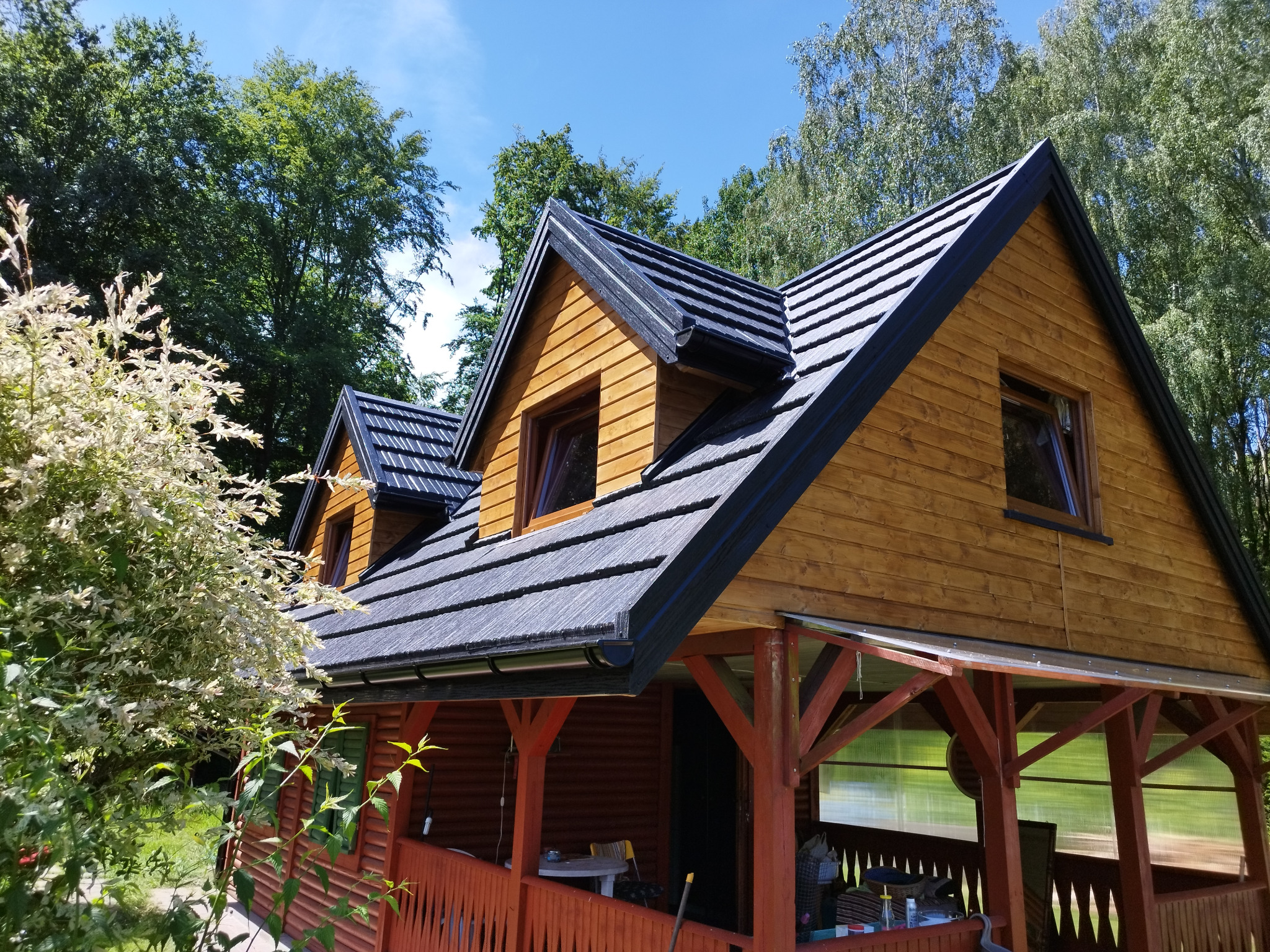 Drewniany domek letniskowy z dwuspadowym dachem pokrytym ciemną dachówką, widok z poziomu ogrodu w słoneczny dzień.
