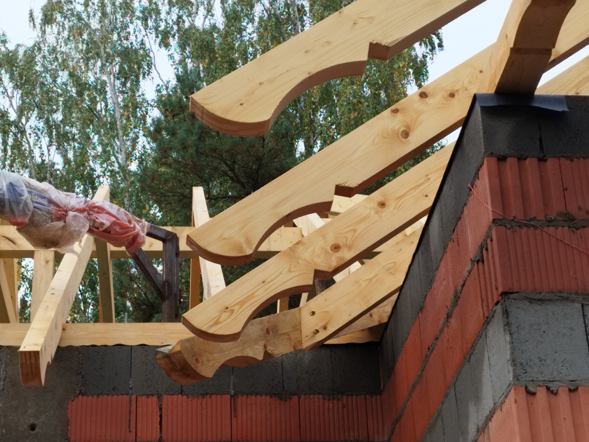 Surowa konstrukcja drewniana z wycięciami na krokwie, wznoszona na murze z czerwonej cegły i szarego betonu, z widocznym fragmentem zieleni drzew w tle.