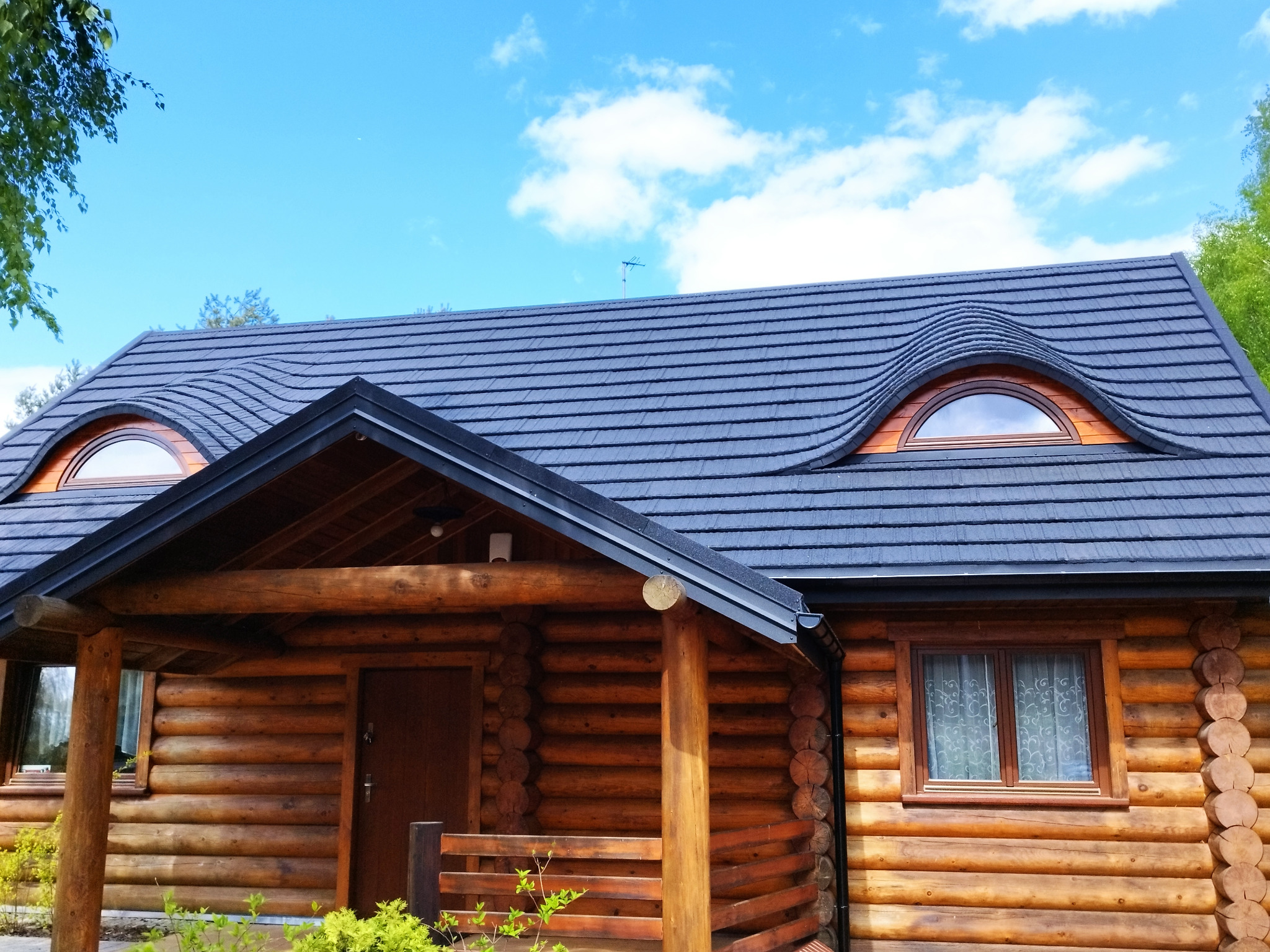 Dom z bali z ciemnoszarym dachem i owalnymi oknami na poddaszu, błękitne niebo w tle.