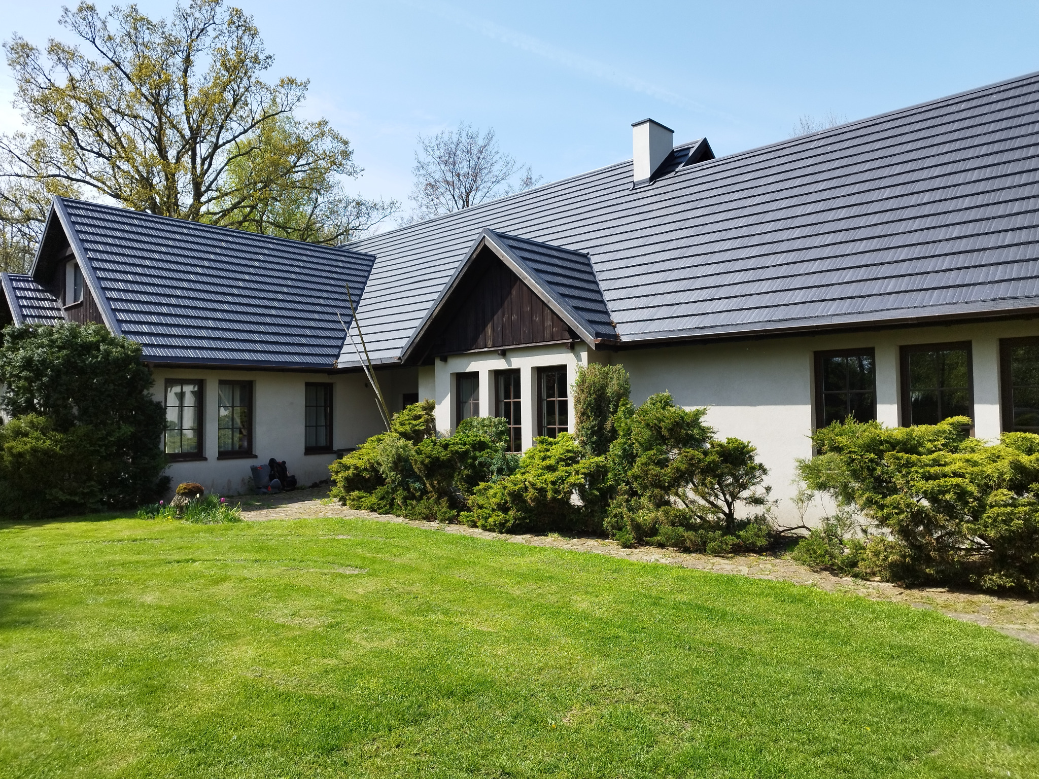 Dom jednorodzinny z ciemnoszarą dachówką ceramiczną, białą elewacją i zadbanym trawnikiem.